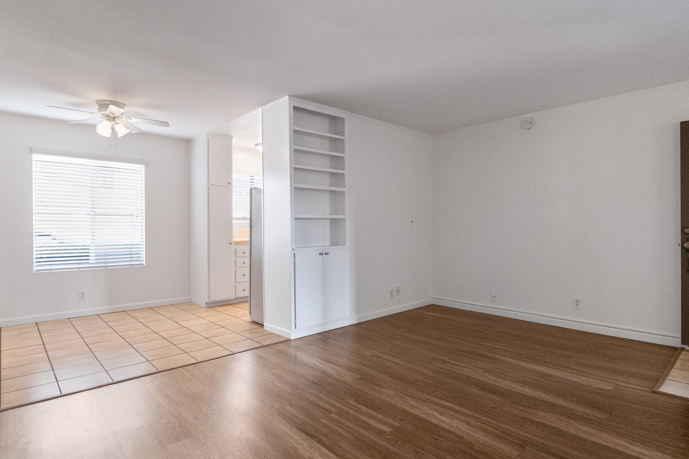 Bright and spacious room featuring a ceiling fan, light wooden floors, and large windows allowing natural light. An open area connects to a tiled floor section, likely a kitchen, with built-in shelves on one side. The walls are painted in a neutral color, creating a clean, inviting atmosphere.