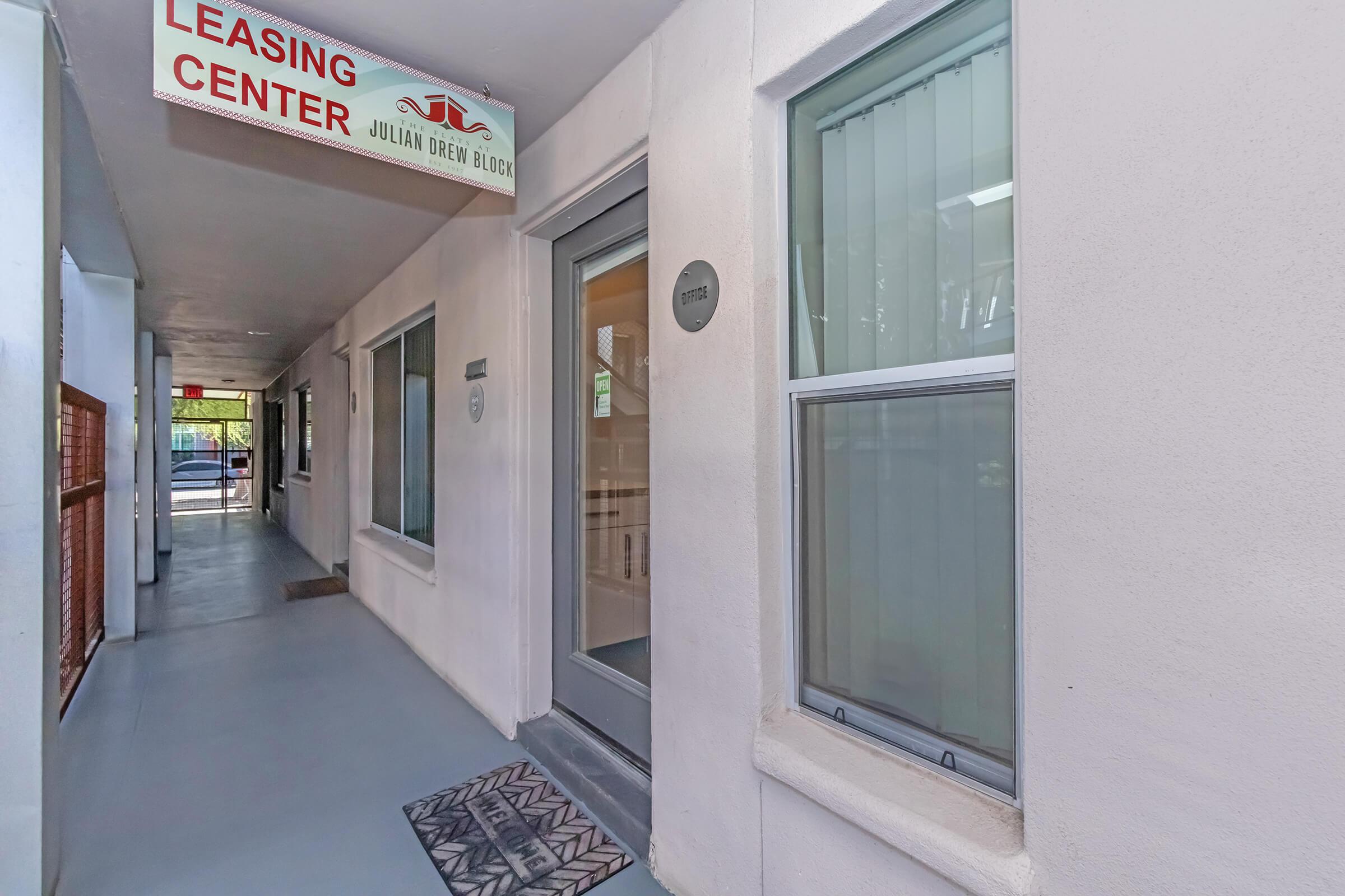 View of a leasing center entrance for the Julian Drew Building, featuring a sign above the door, a doormat in front, and windows on either side. The hallway is well-lit with a smooth floor leading to an outdoor area.