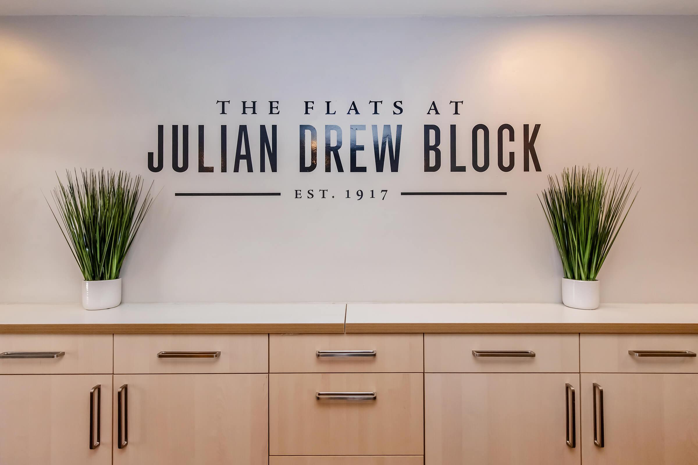 Wall with the text "The Flats at Julian Drew Block" and "Est. 1917" in bold letters. There are two potted plants on either side of the text, placed on a light wood counter. The overall color scheme is neutral with a modern design.