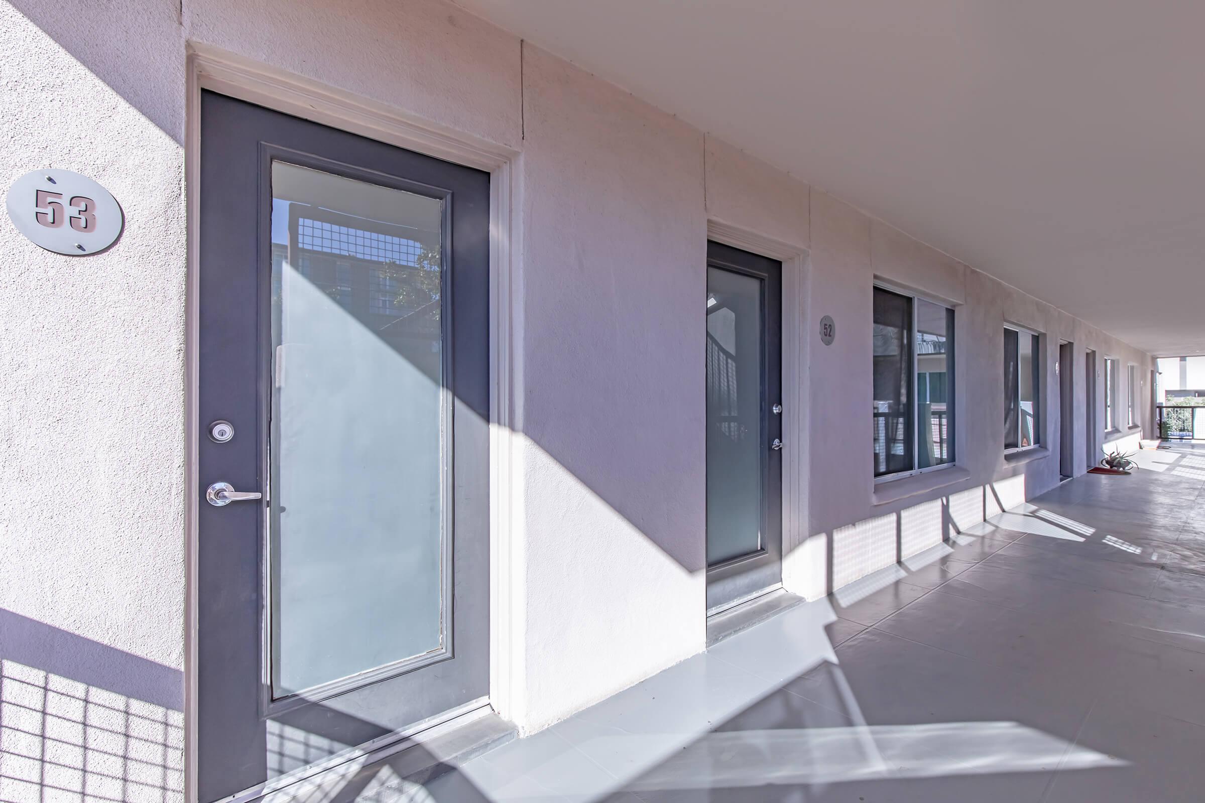 A row of modern apartment doors with large windows, featuring light-colored walls and a numbered door marked "53." Soft shadows are cast on the ground, indicating bright sunlight. The entrance area is clean and well-maintained, contributing to a welcoming atmosphere.