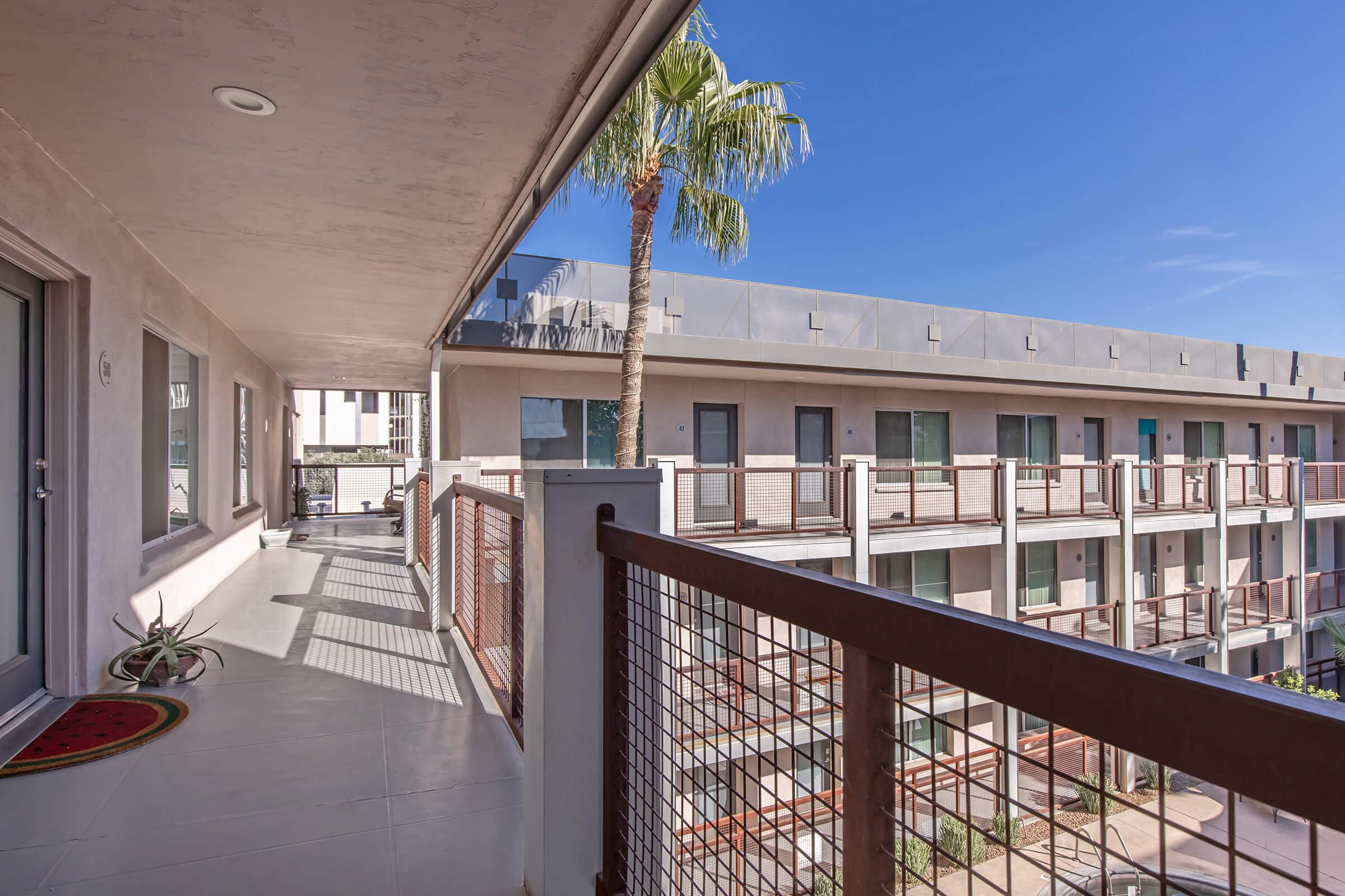 A bright outdoor balcony with a palm tree and a decorative mat, overlooking a modern apartment building with multiple units. The architecture features a mix of beige and gray tones, with large windows allowing natural light. The clear blue sky adds to the overall vibrant atmosphere.