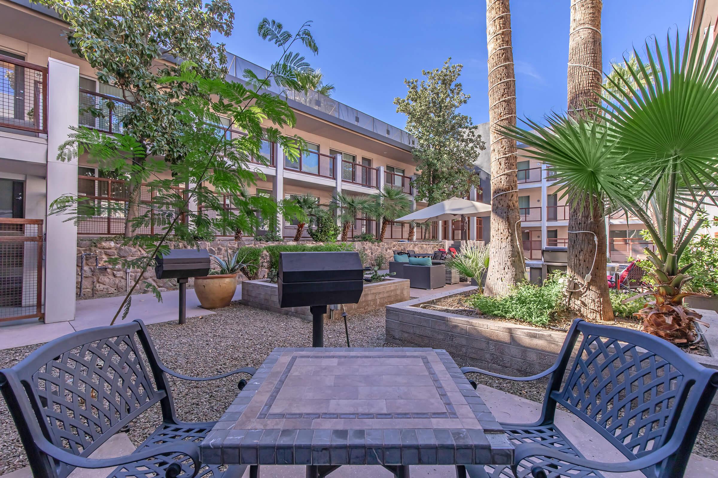 A sunny courtyard featuring potted palm trees, two outdoor grills, and a cozy seating area with a table and chairs. The surroundings include landscaped greenery and modern architecture, creating an inviting atmosphere for relaxation and socializing.
