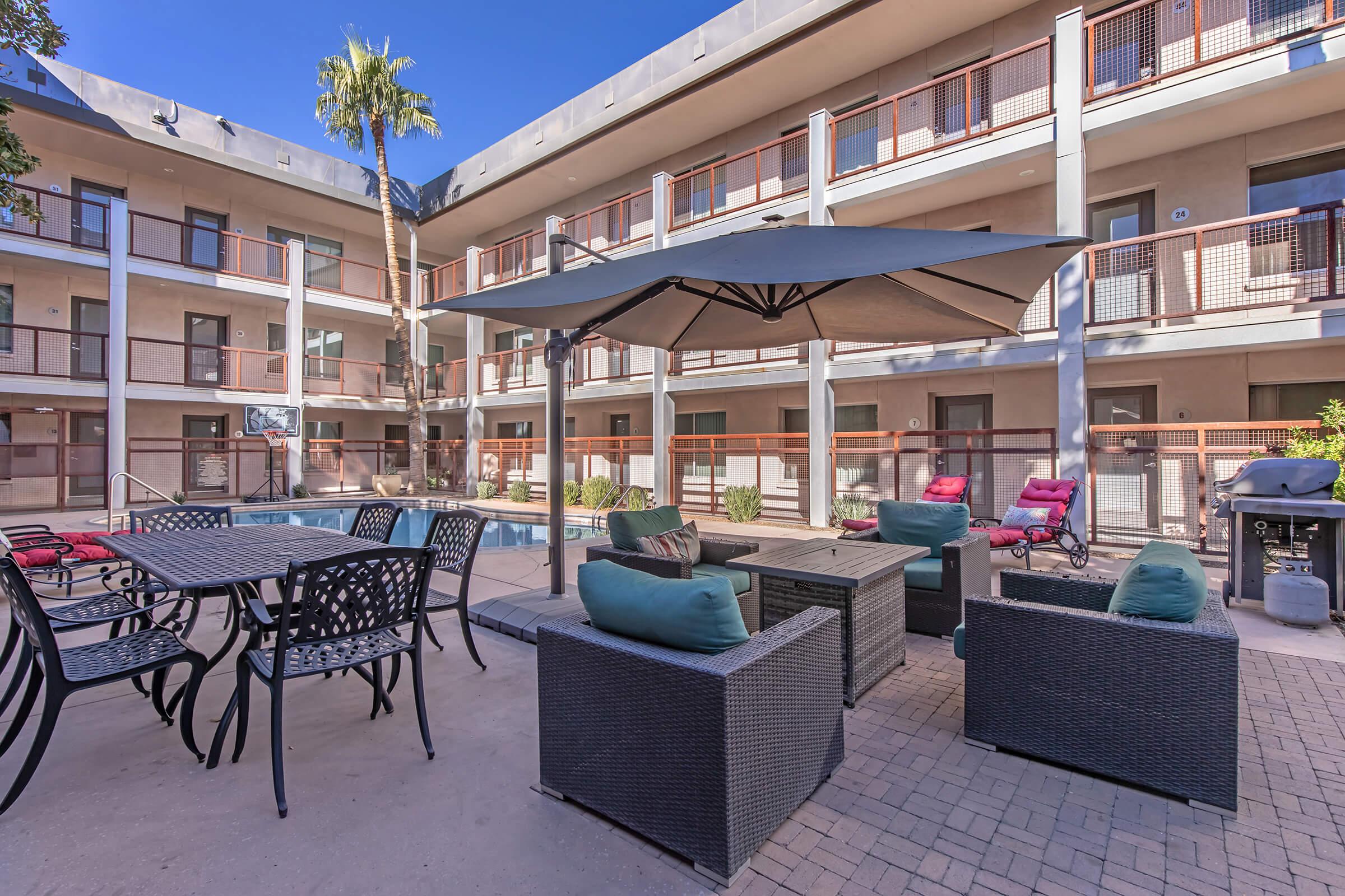 An outdoor area featuring a swimming pool surrounded by a patio space. There are comfortable seating arrangements with cushions, a table with chairs, and an umbrella for shade. The background includes two-story building facades with balconies and a palm tree.
