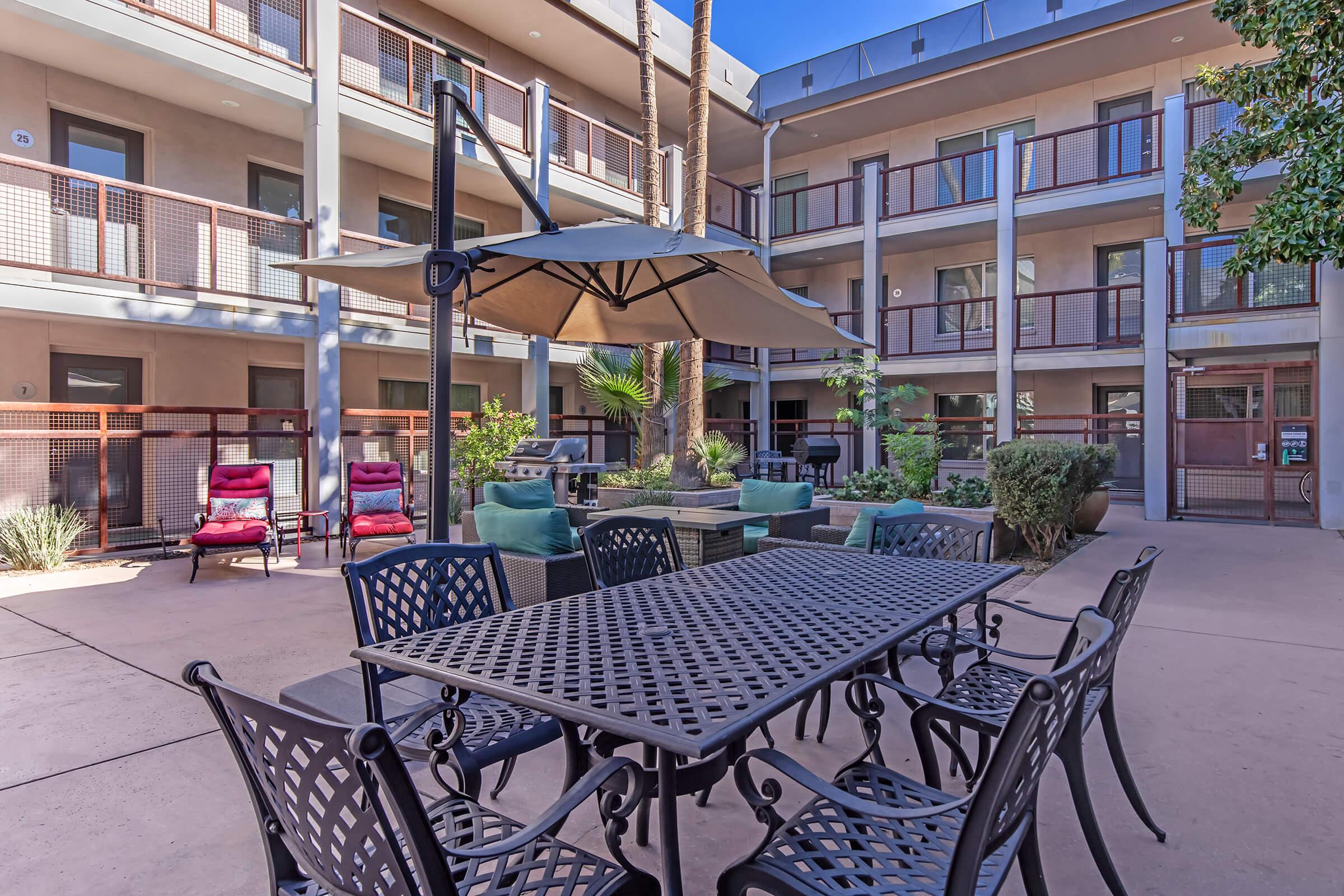 A courtyard with a shaded table and chairs surrounded by lush greenery and potted plants. There are comfortable seating areas with sofas and armchairs, and a few palm trees provide a tropical ambiance. The background features modern apartment buildings with balconies.