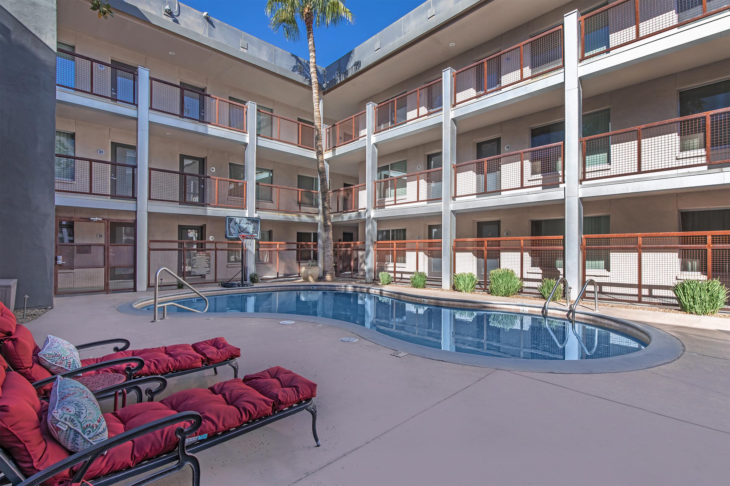 A view of a courtyard with a swimming pool surrounded by a few palm trees. There are lounge chairs with red cushions arranged near the pool. The building features multiple levels with balconies and a modern aesthetic, and the area is well-lit by sunlight.