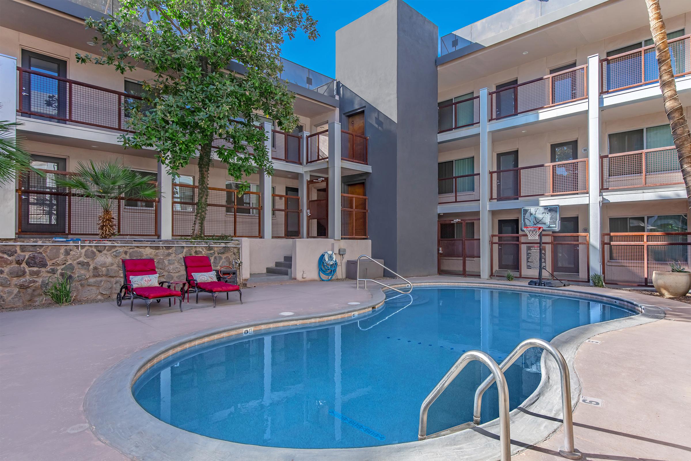 A tranquil courtyard featuring a swimming pool surrounded by palm trees and two red lounge chairs. The area is enclosed by a modern building with balconies, showcasing a blend of natural and architectural elements under a clear blue sky.