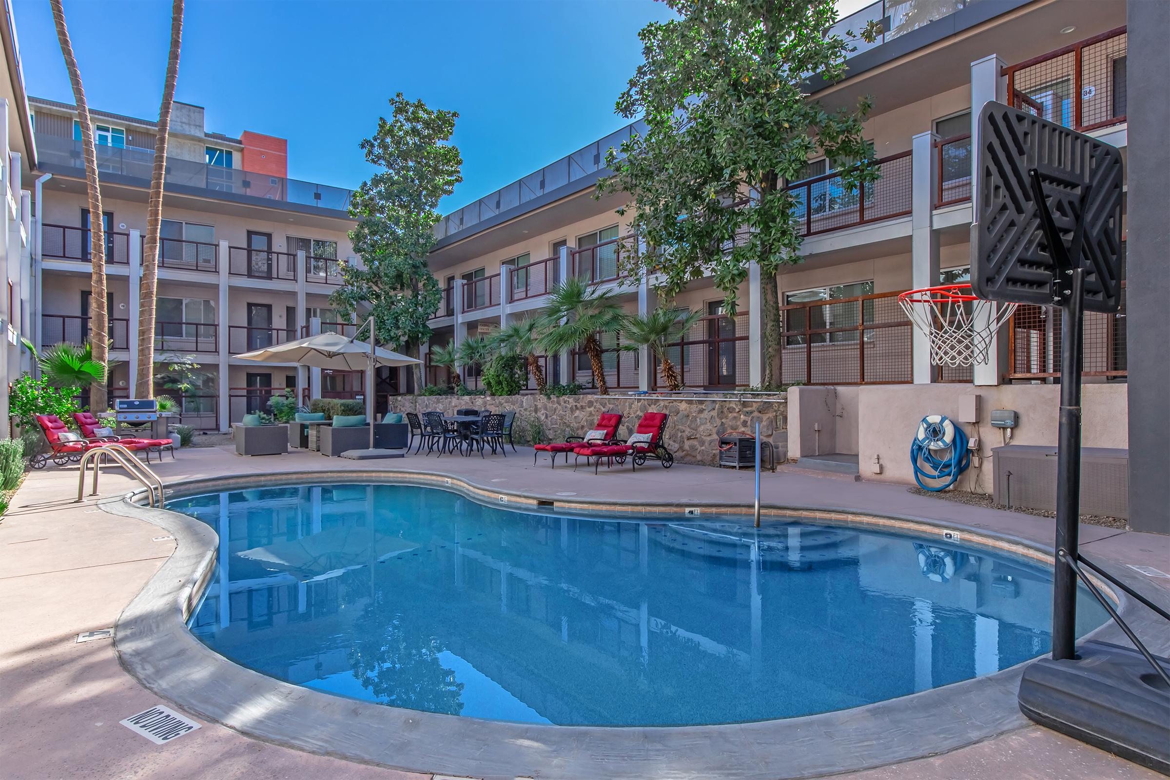 A tranquil swimming pool surrounded by palm trees and lounge chairs, situated in a courtyard of an apartment complex. A basketball hoop is positioned nearby, and outdoor seating with tables and umbrellas is visible, creating a relaxing atmosphere for residents and guests.