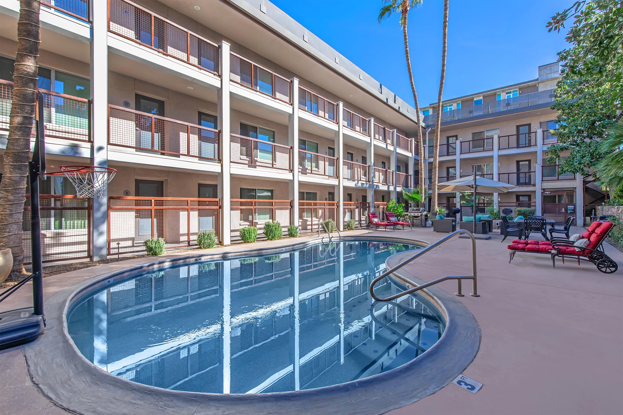 A peaceful hotel courtyard featuring a small round swimming pool surrounded by lounge chairs. The area is adorned with palm trees and greenery, with balconies overlooking the pool. A basketball hoop is visible, adding to the recreational atmosphere. Bright blue sky enhances the relaxing environment.
