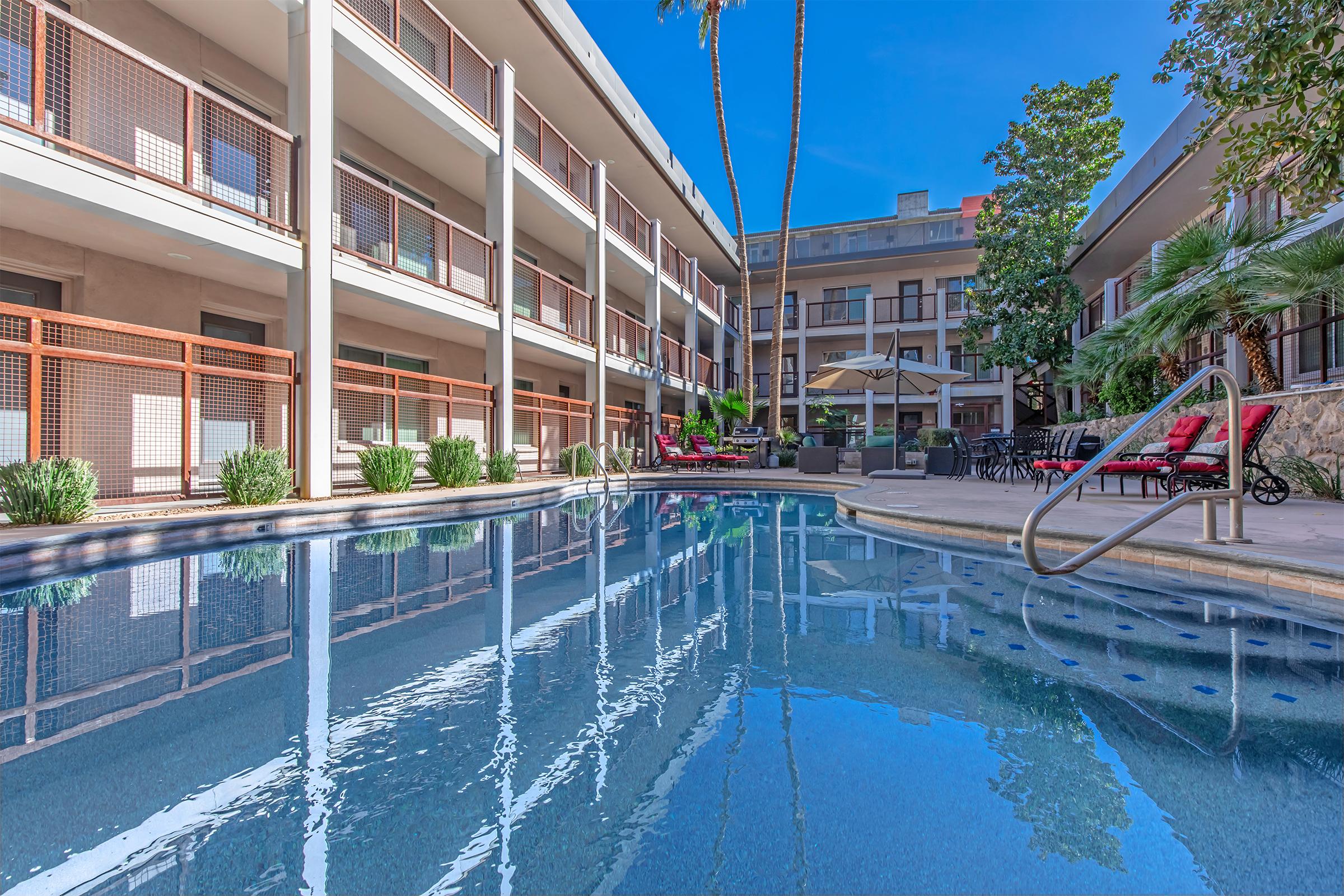 A clear blue swimming pool reflecting the sky, bordered by lush green plants. Surrounding the pool are modern buildings with balconies, shaded seating areas, and palm trees under a bright, sunny sky. The atmosphere exudes relaxation and hospitality.