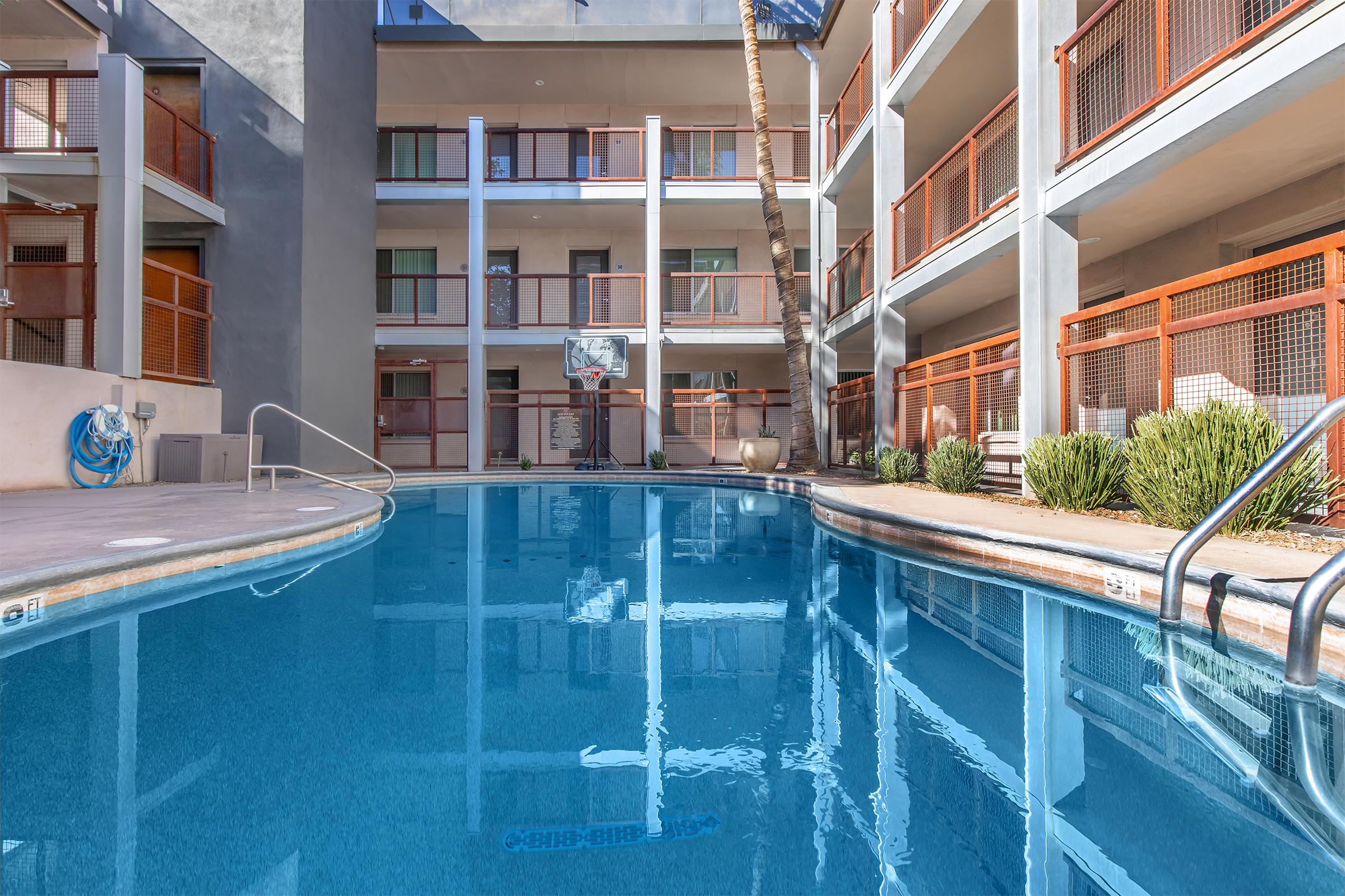 A clear pool reflects the surrounding buildings and palm trees in a modern courtyard. The pool area features a slide, lounge chairs, and nearby balconies with orange railings. The setting is bright and inviting, showcasing a relaxing atmosphere for guests.