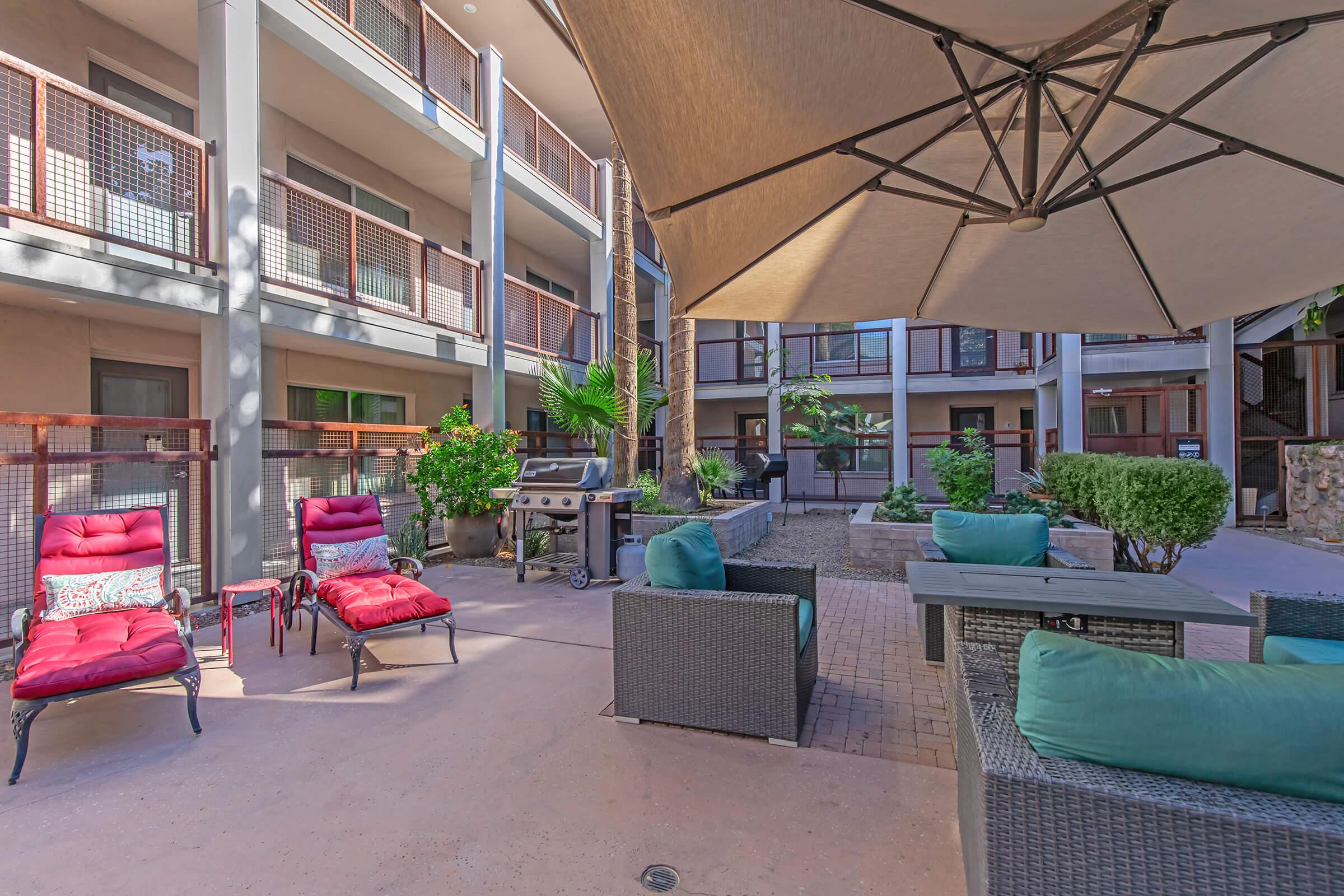 A cozy outdoor lounge area featuring comfortable seating with colorful cushions, a barbecue grill, and lush greenery. Shade is provided by a large umbrella, and the surroundings include modern apartment balconies, creating an inviting atmosphere for relaxation and socializing.