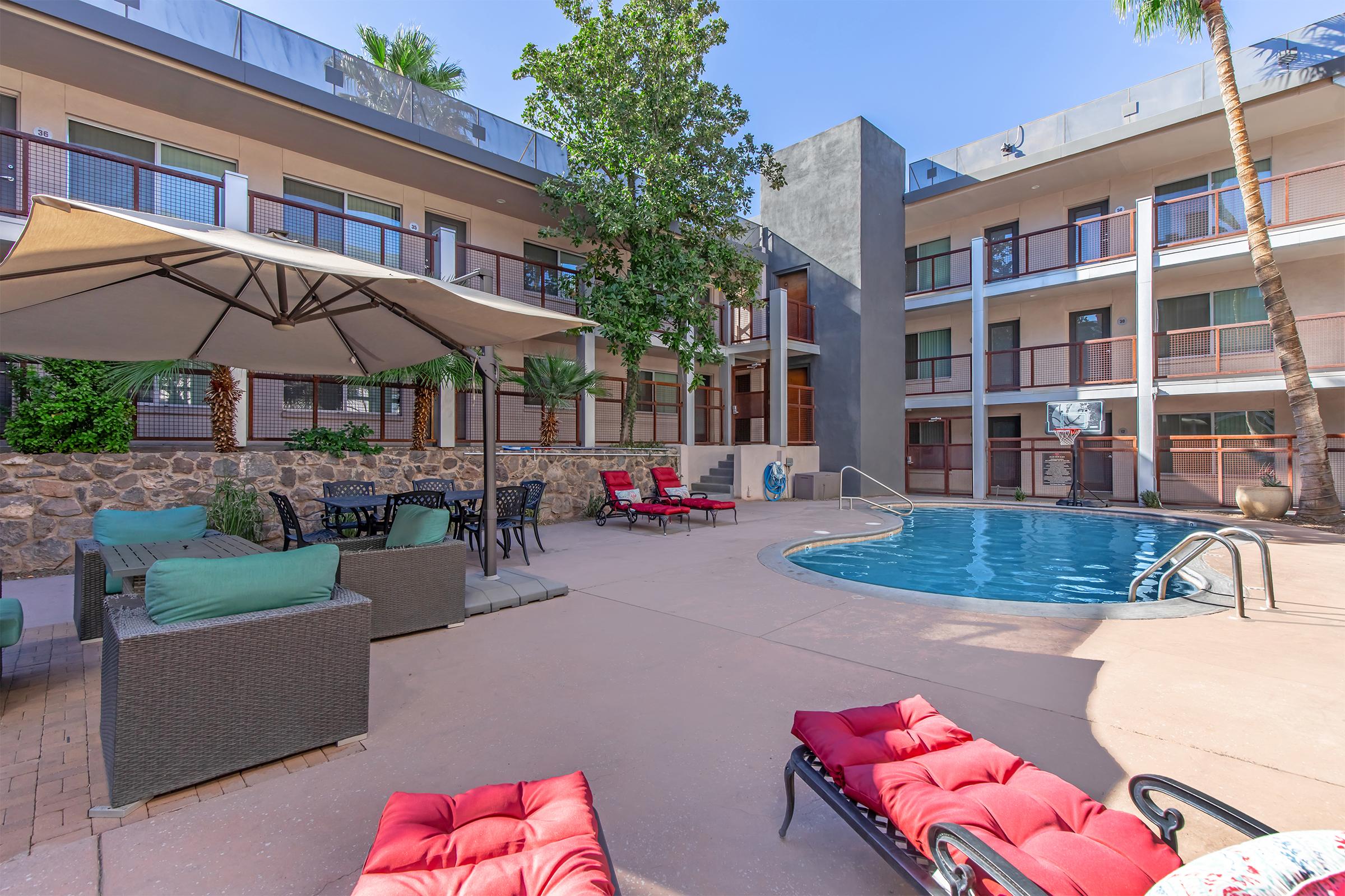 A sunny outdoor space featuring a swimming pool surrounded by lounge chairs and a shaded seating area. Palm trees and greenery enhance the relaxing atmosphere, with a building featuring balconies in the background. A basketball hoop is visible nearby, adding to the recreational options.