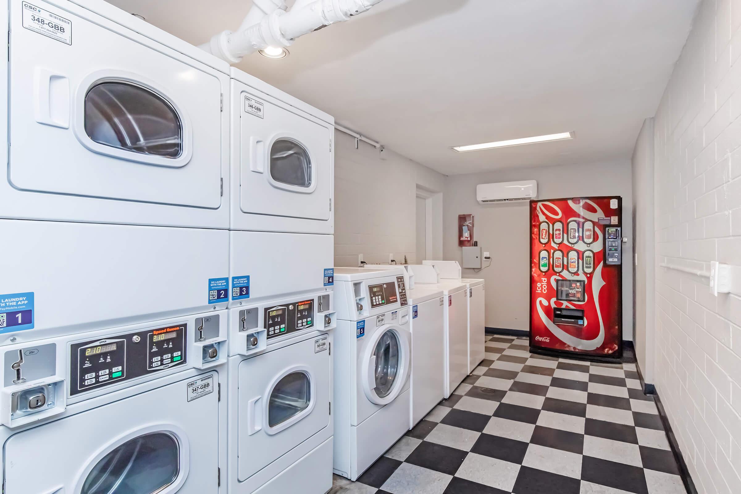 A clean laundry room featuring several stacked washing machines and dryers with digital control panels, alongside a vending machine against a white wall. The floor is checkered in black and white tiles. Bright lighting illuminates the space, making it inviting for patrons.