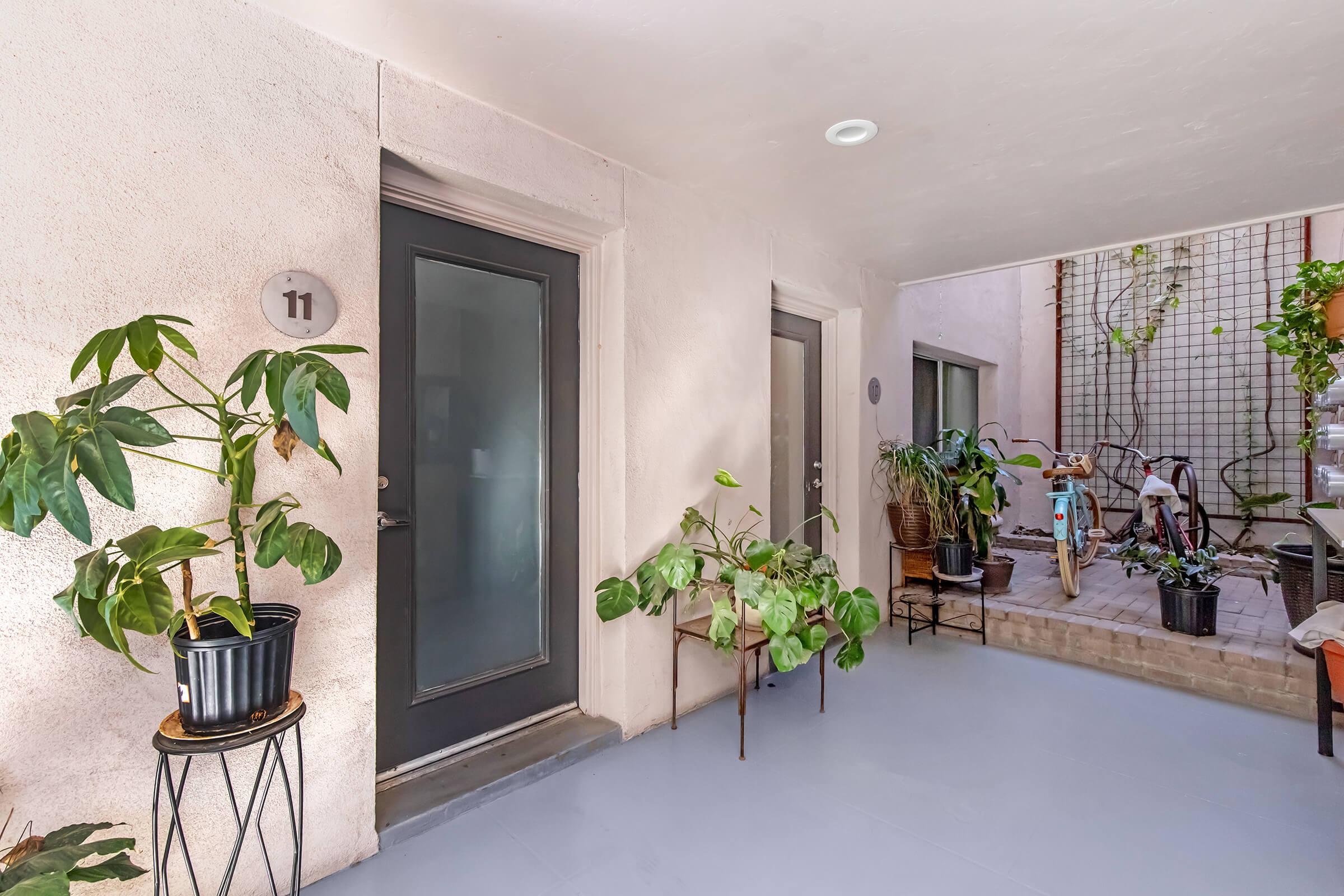 A well-lit entryway featuring two doors, with the number 11 prominently displayed. Potted plants are positioned on either side, adding a touch of greenery. The space is clean and minimalistic, with a bicycle and additional plants visible in the background, creating a welcoming atmosphere.
