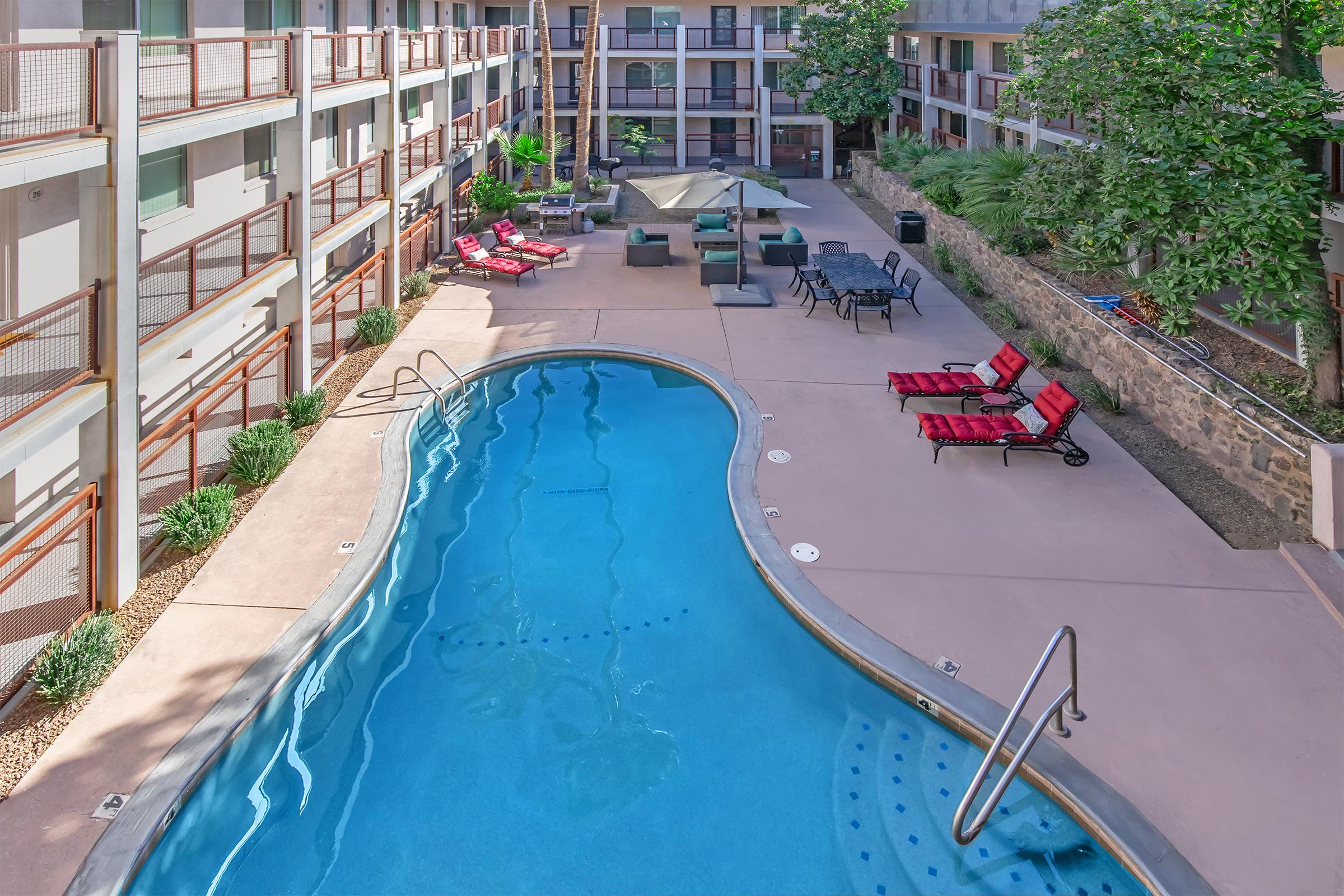 Aerial view of a hotel courtyard featuring a blue swimming pool surrounded by lounge chairs and patio furniture. Lush greenery is visible in the background alongside a stone wall, with additional seating areas and umbrellas enhancing the outdoor space. The scene is bright and inviting, reflecting a relaxing atmosphere.