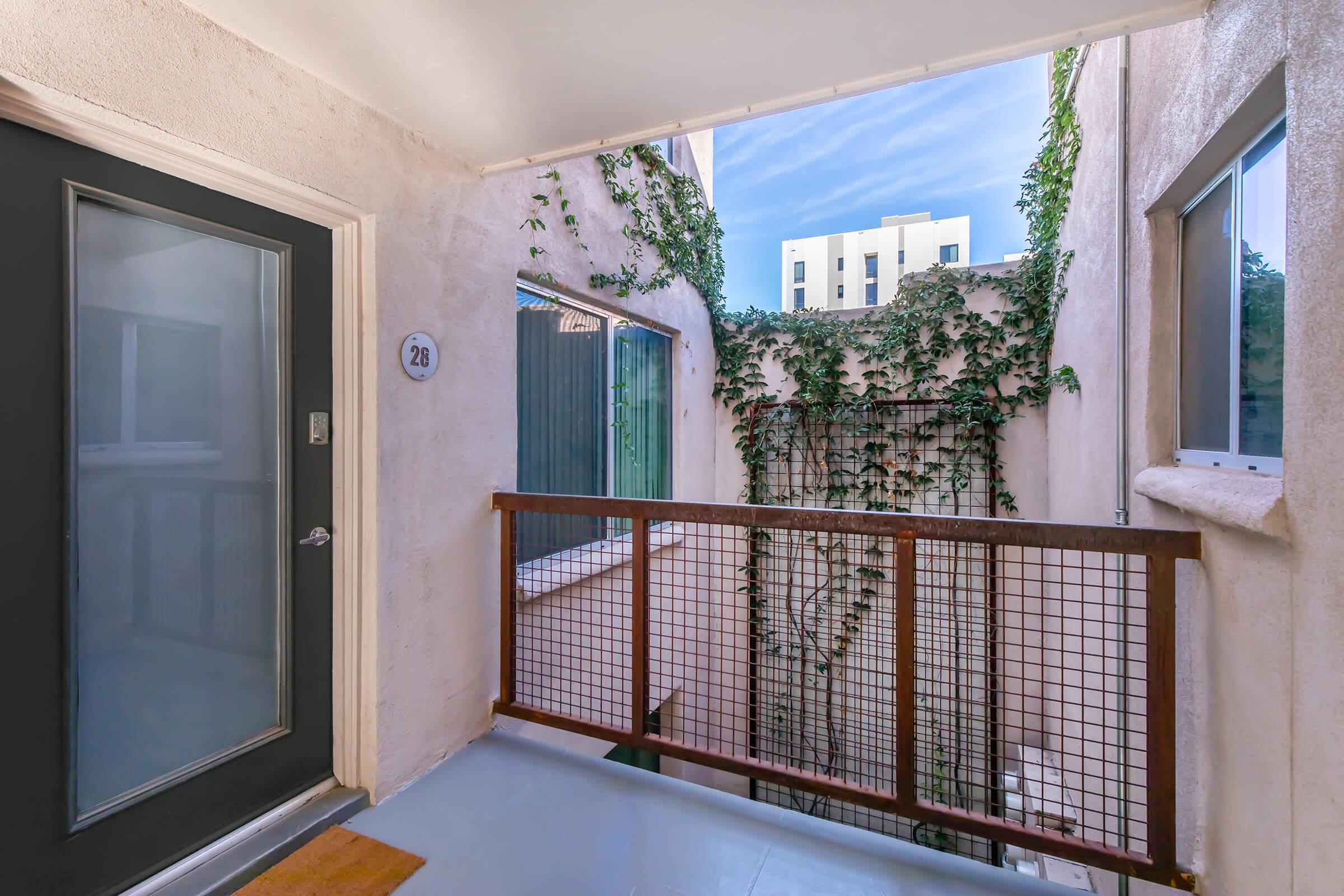 A view from a balcony or corridor with a dark door labeled "26." The space is surrounded by light-colored walls and features greenery climbing the walls. There are windows visible on either side, with a clear blue sky above. The area appears bright and inviting.