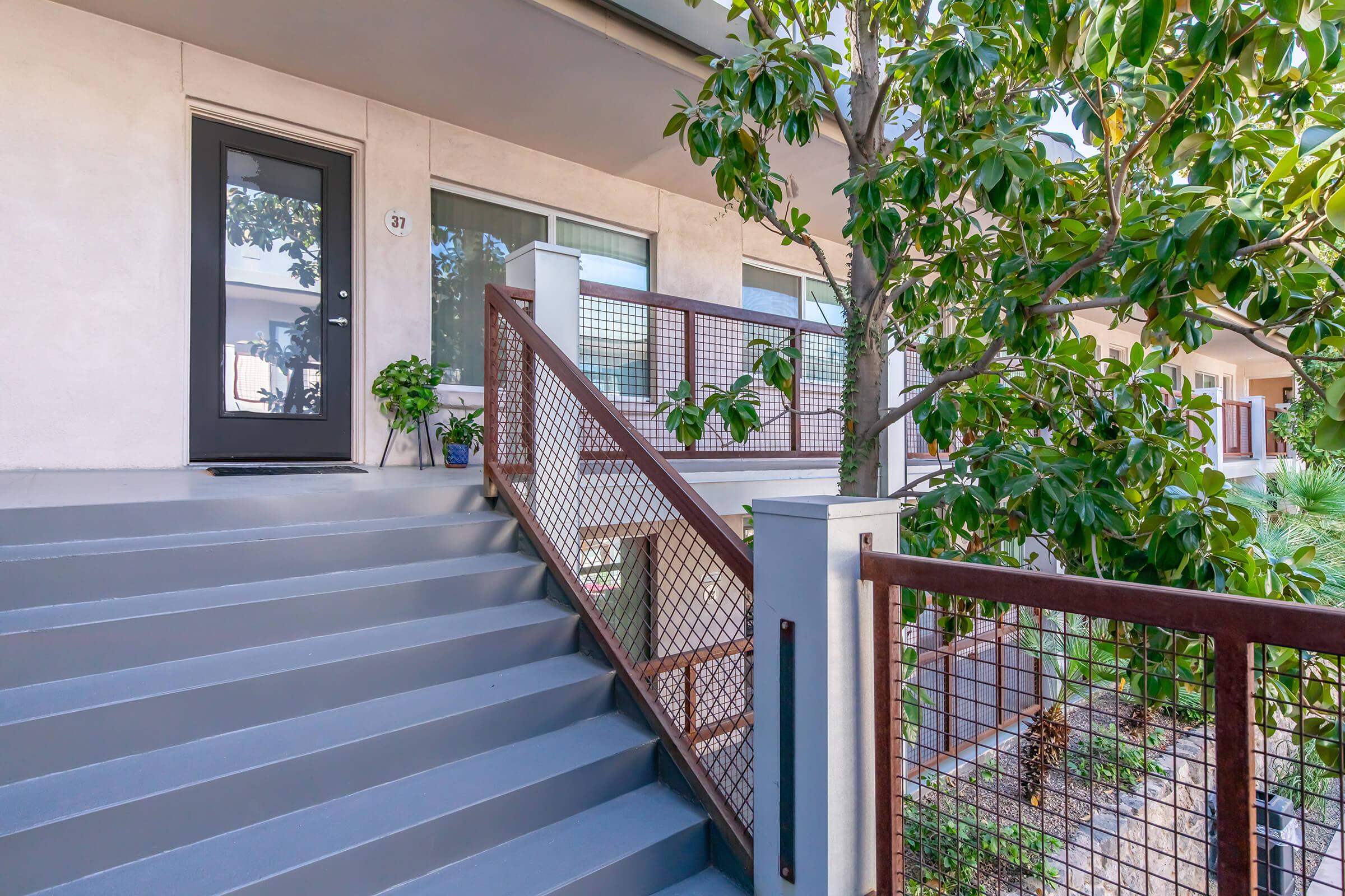 A modern entrance featuring a black door and a set of stairs leading up to it. The staircase has a metal railing and is bordered by green plants. In the background, there are windows visible, and a tree adds greenery to the scene. The overall aesthetic is clean and contemporary.