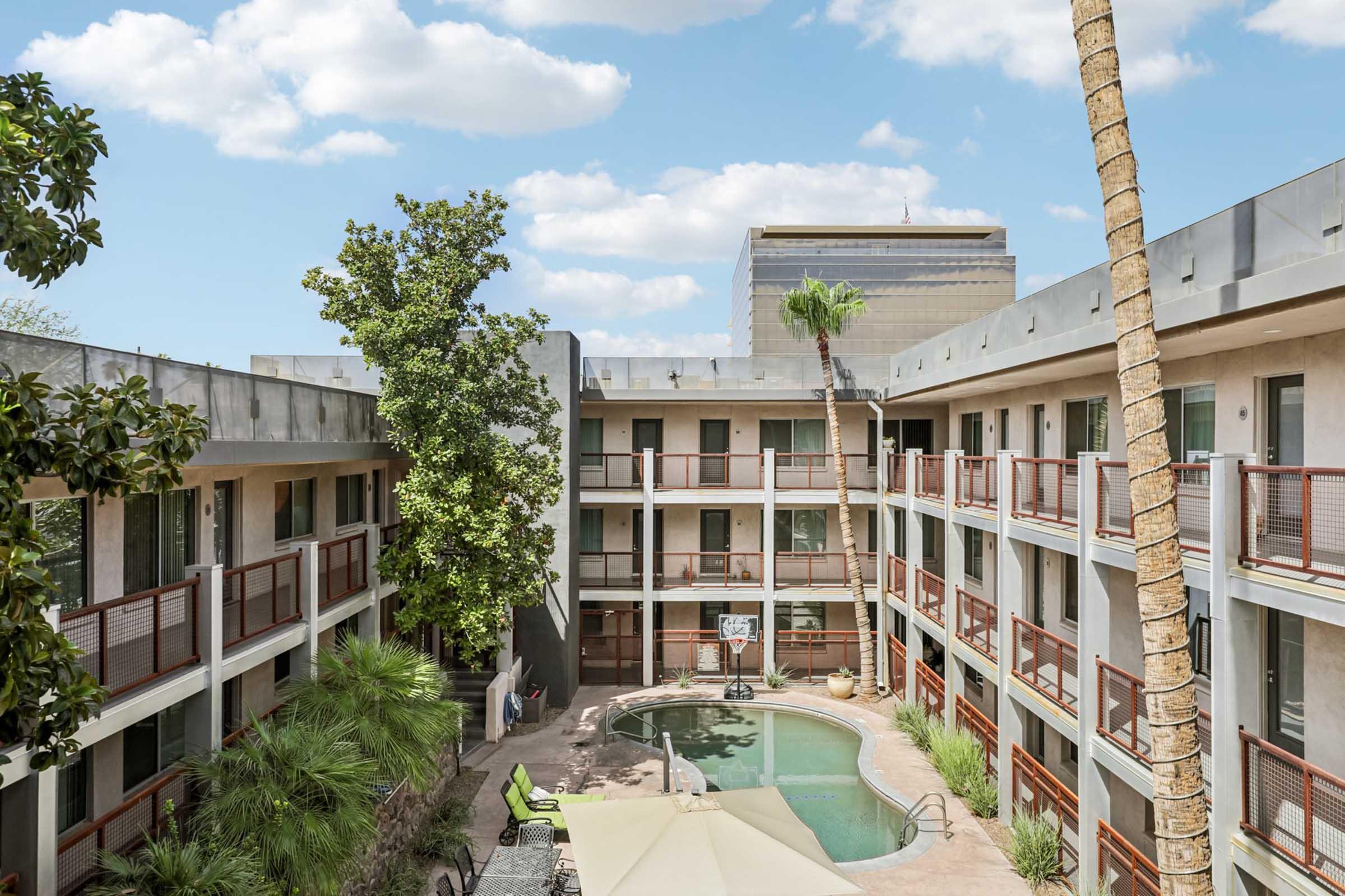 A view of a modern hotel courtyard featuring a swimming pool surrounded by palm trees and greenery. The hotel has a contemporary design with multiple levels and balconies. Bright blue skies with a few clouds overhead enhance the inviting atmosphere.