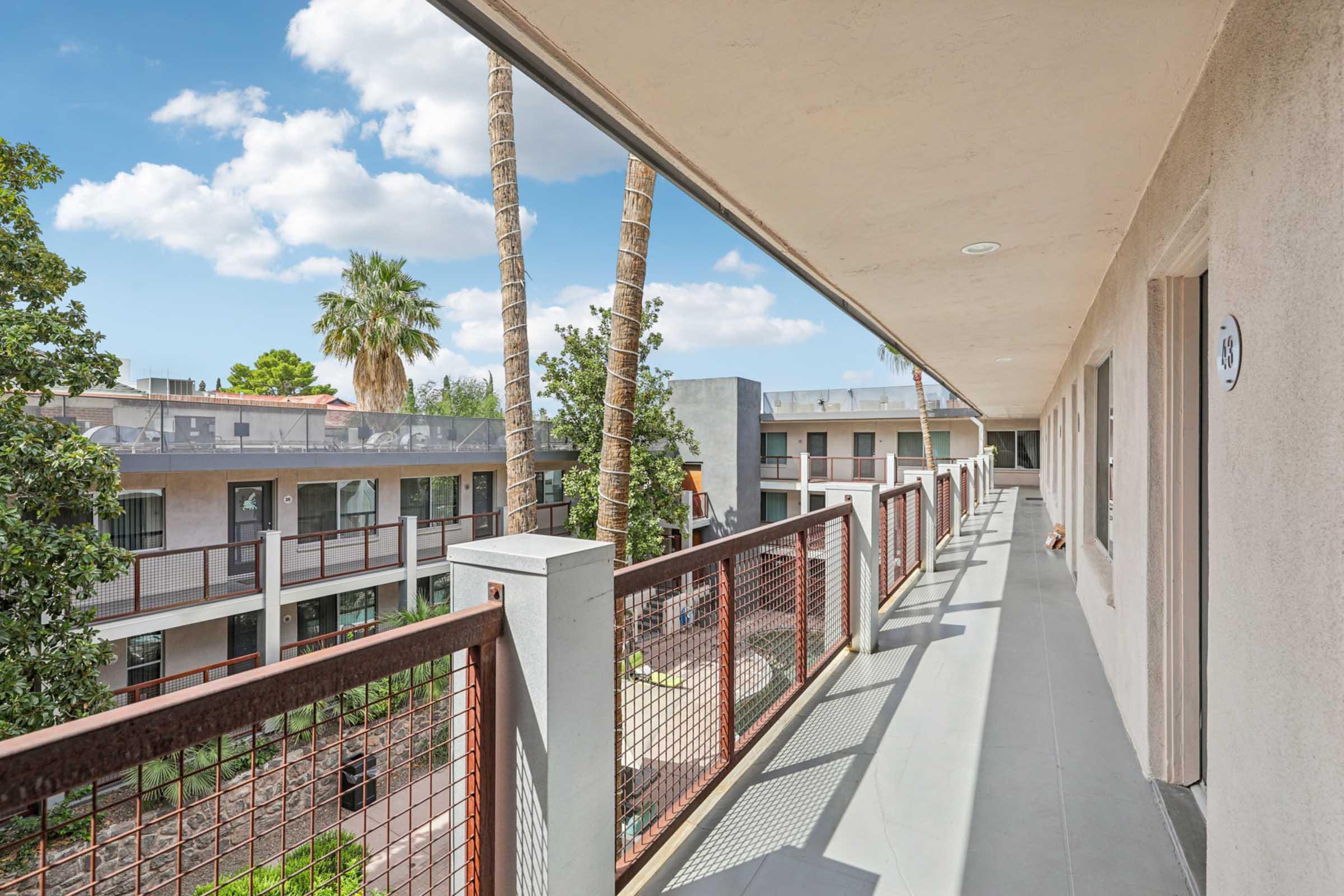 A modern hotel balcony view showing palm trees and a courtyard. The walkway has a sleek railing, and the building features contemporary architecture with large windows and outdoor spaces. A partly cloudy blue sky enhances the bright and inviting atmosphere.