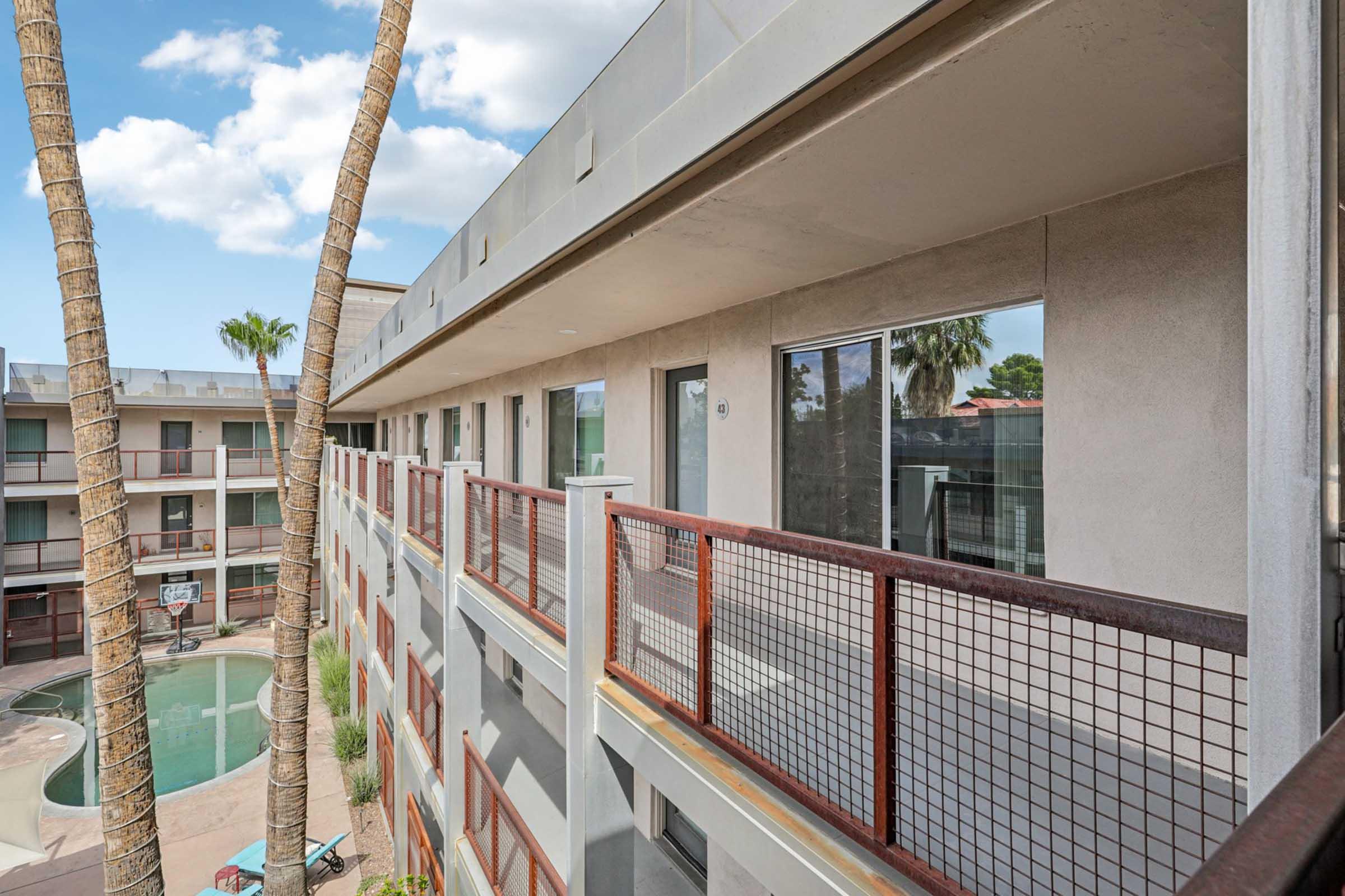 A view of a hotel balcony overlooking a courtyard with a swimming pool, surrounded by tall palm trees. The building features multiple levels, with glass doors leading to guest rooms. The sky is partly cloudy, adding to the relaxed atmosphere of the setting.