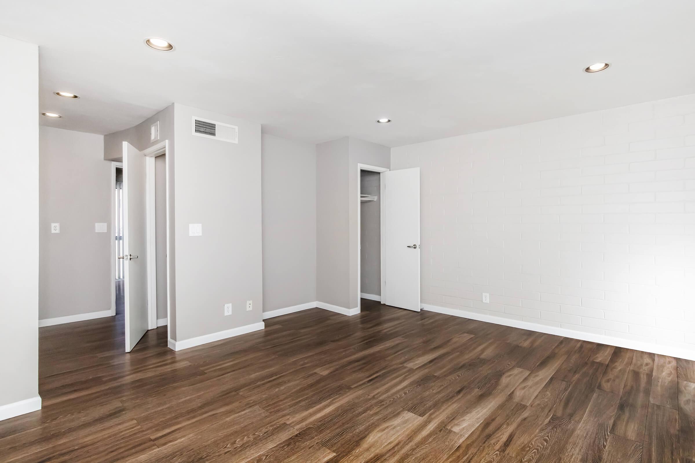 Empty modern room with light gray walls and dark wood flooring. A white interior door leads to a closet, and another door is partially visible in the background. The space has recessed lighting on the ceiling, giving it a bright and clean look.
