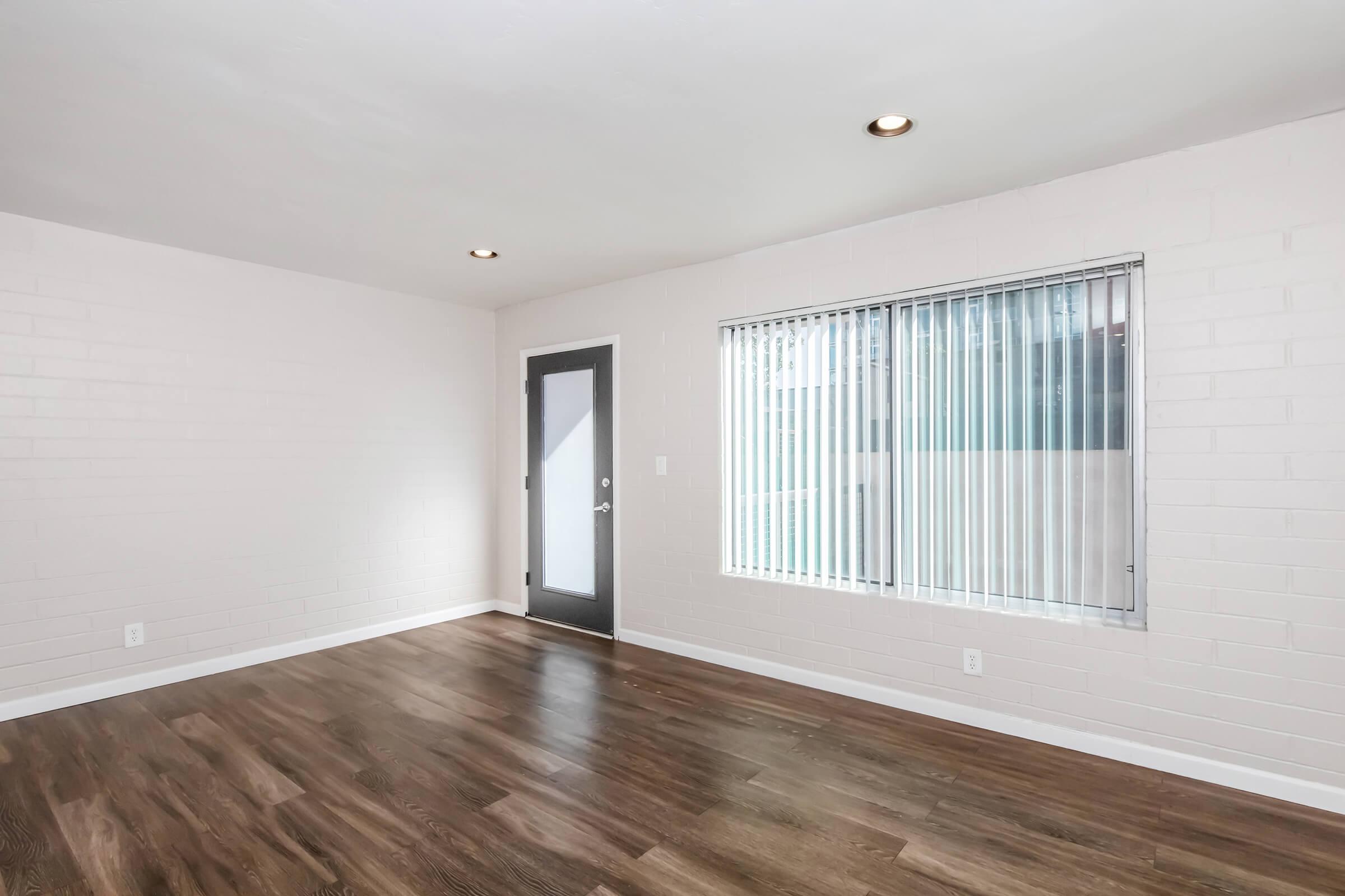 Empty room featuring light-colored brick walls, a large window with vertical blinds, and a dark door. The floor is made of dark wood. Natural light is coming in through the window, creating a bright atmosphere.