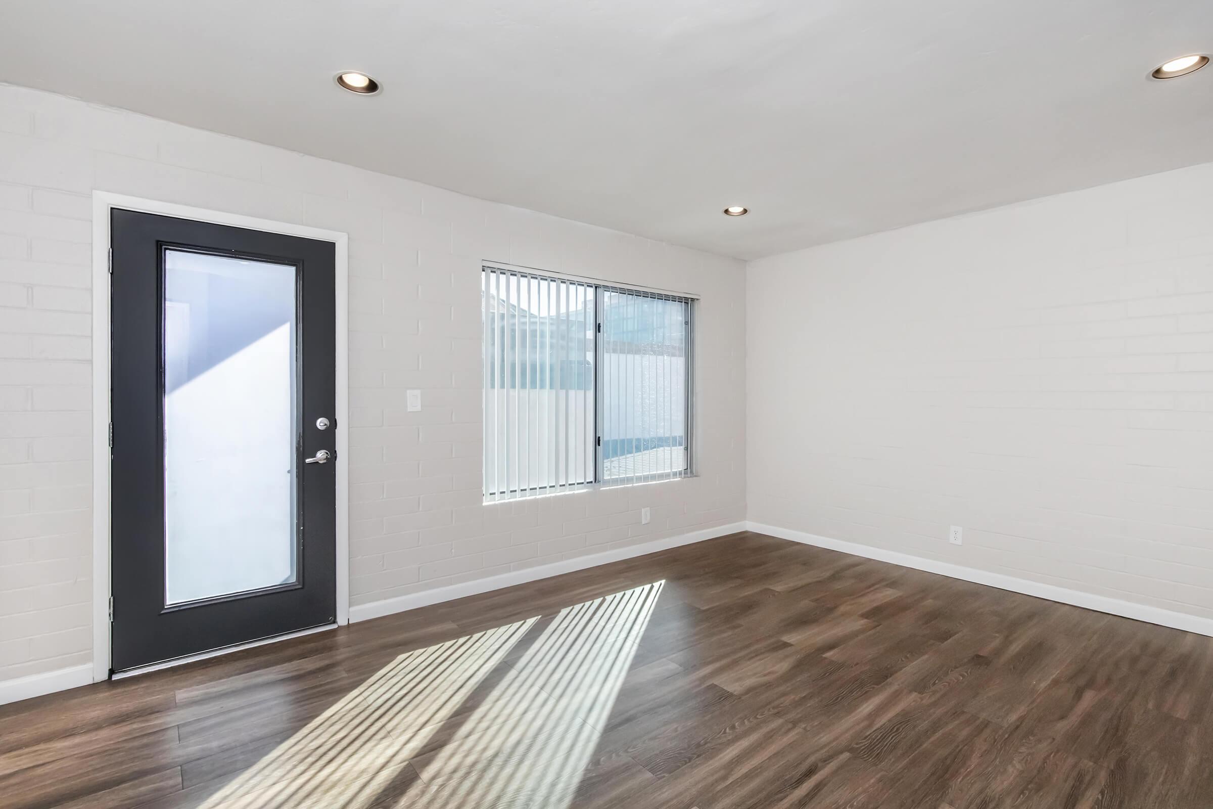 Bright, empty room featuring a large window with blinds, a black front door, and light hardwood flooring. The walls are painted white, creating a clean and spacious atmosphere. Natural light streams in through the window, casting shadows on the floor.