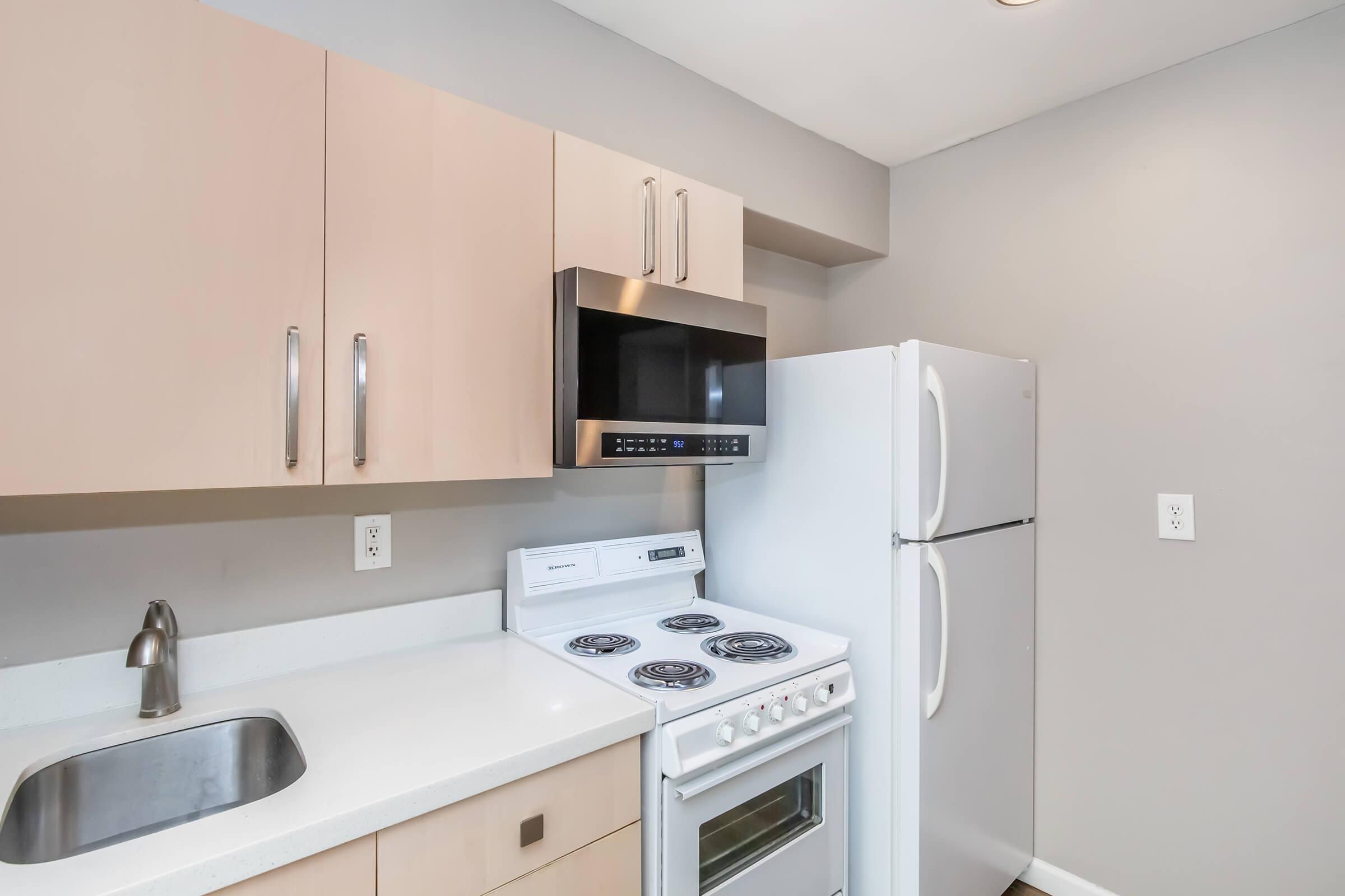 Modern kitchen featuring light-colored cabinetry, a stainless steel microwave, a white refrigerator, and a white stove with an oven. The countertop is light and smooth, with a small stainless steel sink. The walls are painted in a neutral gray color, creating a clean and contemporary look.