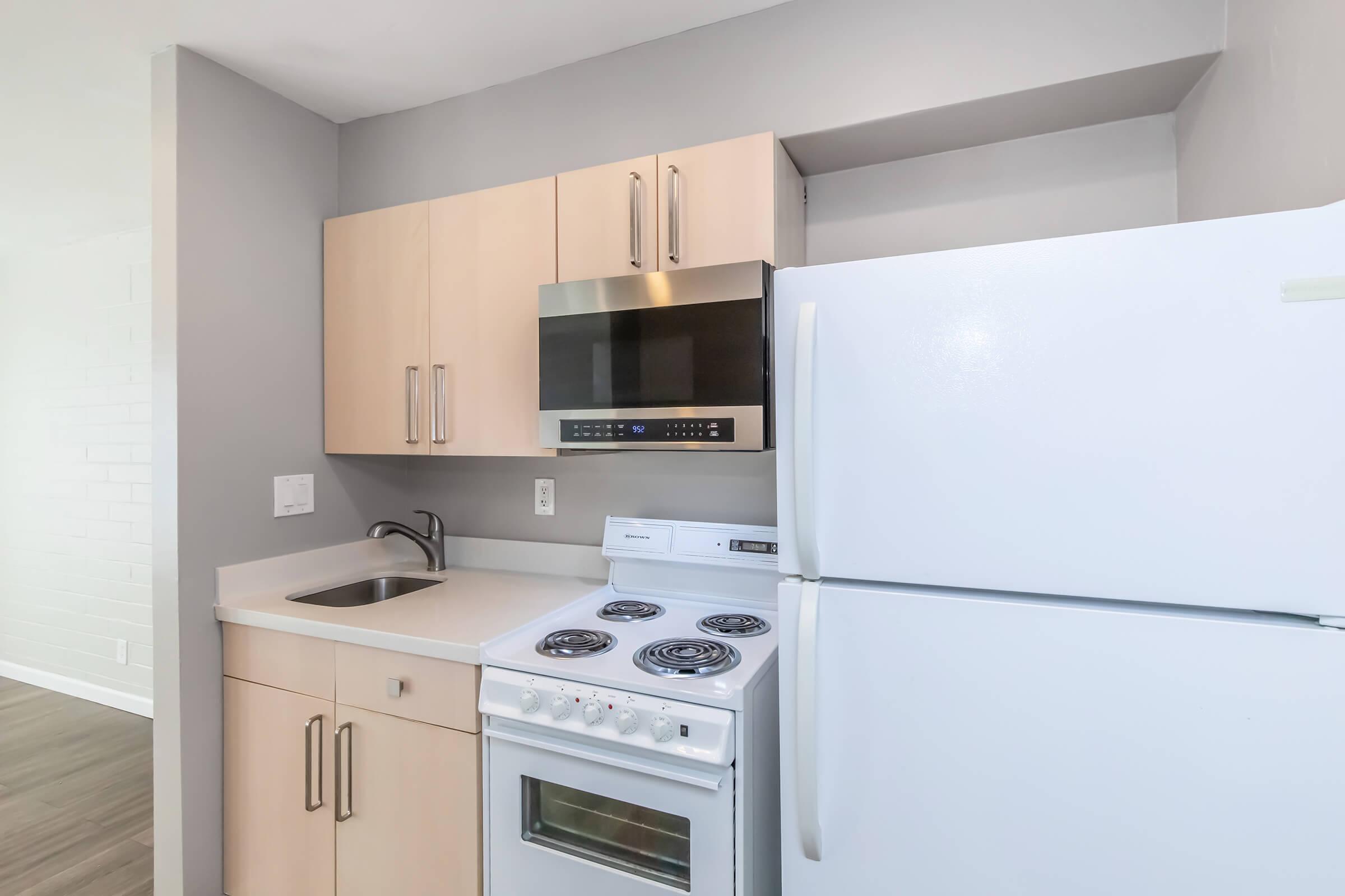A modern kitchen interior featuring light wood cabinetry, a white stove with an oven, a microwave mounted above, a stainless steel sink, and a white refrigerator. The walls are painted in a soft gray color, and the flooring appears to be wood. Natural light fills the space, creating an inviting atmosphere.