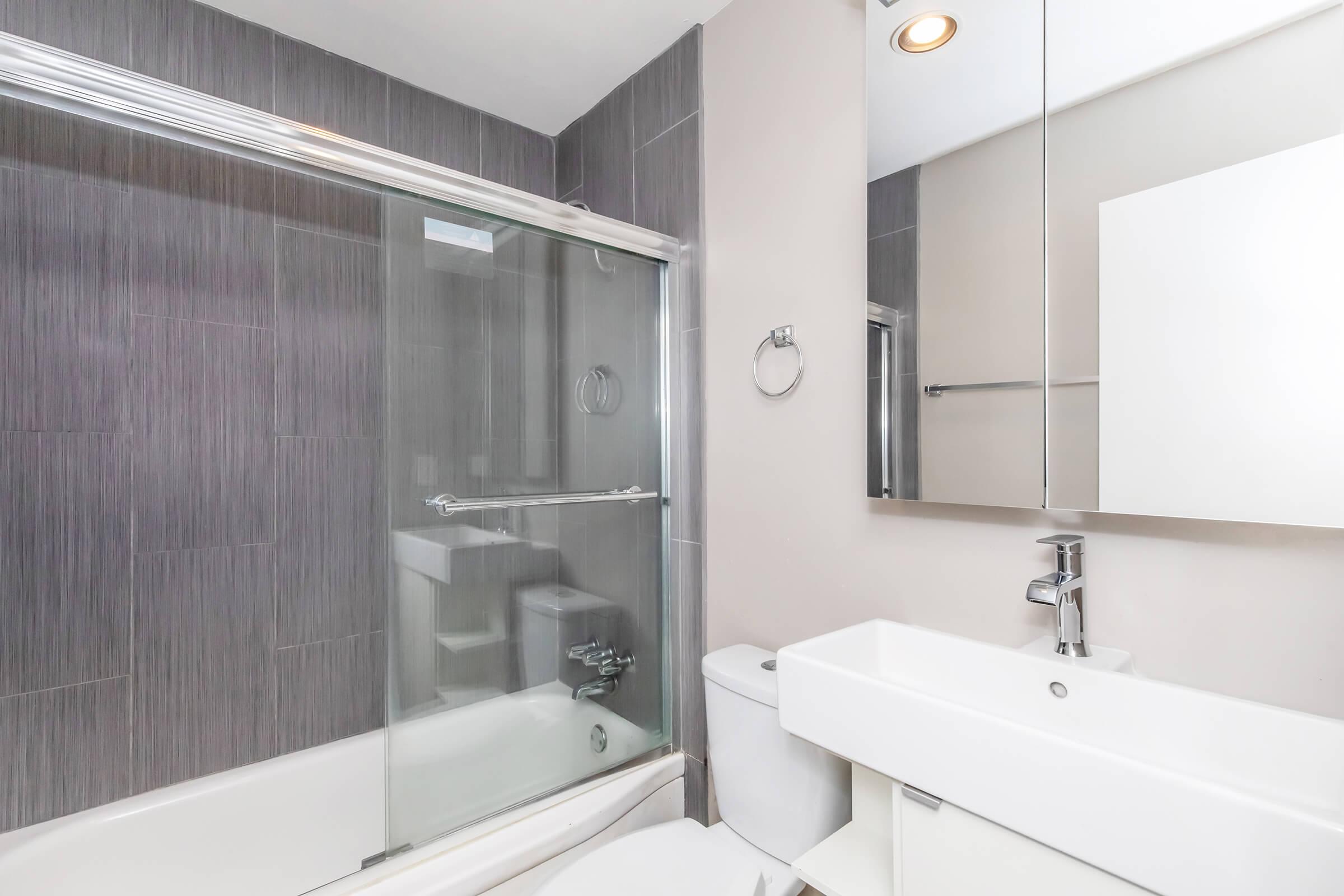 Modern bathroom featuring a glass shower enclosure, sleek bathtub, wall-mounted sink with a minimalist design, and a large mirror. The walls are tiled in a dark gray color, and there is a silver towel ring and a toilet in the corner, creating a clean and contemporary aesthetic.