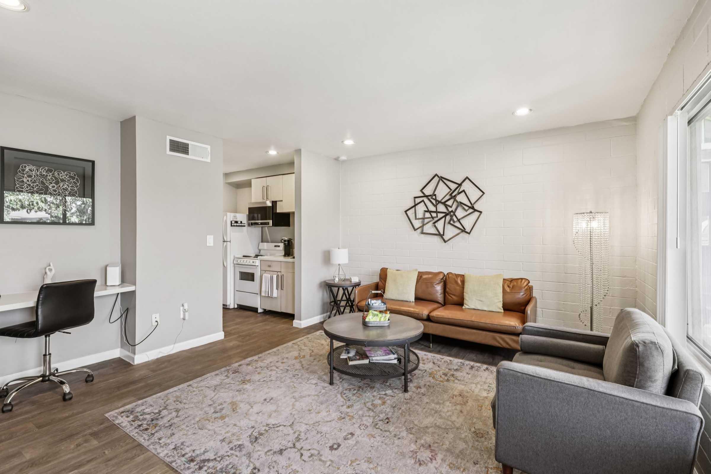 A modern living room featuring a leather sofa, a gray armchair, and a round coffee table on a decorative rug. A wall art piece is displayed behind the sofa, and a small workspace with a desk is visible. Bright lighting and an open kitchen area create a spacious and inviting atmosphere.