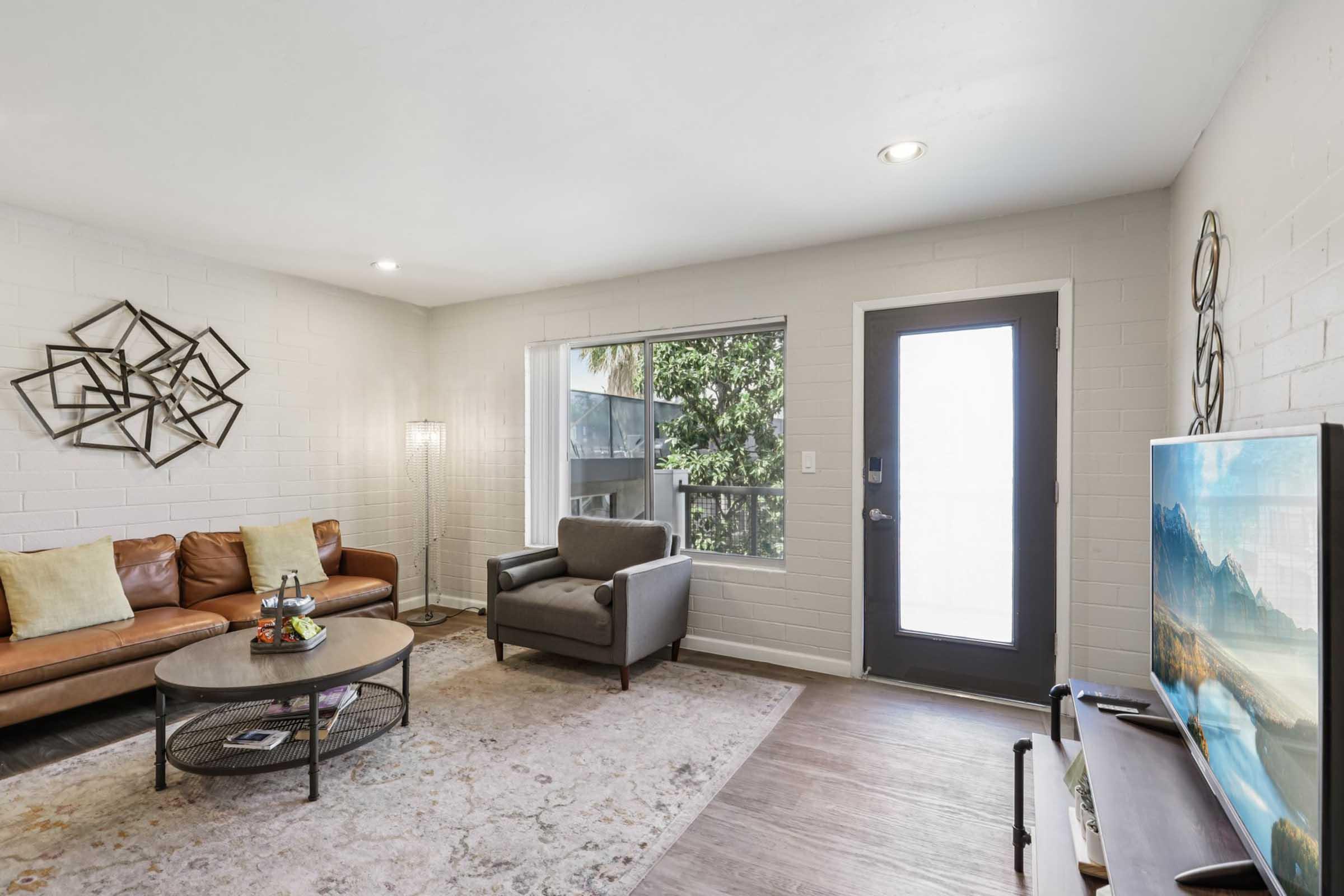 A modern living room featuring a brown leather sofa with decorative pillows, a round coffee table, and a grey armchair. There's a large window allowing natural light, a door leading outside, and a TV on a stand. The room has a neutral color palette with a subtle area rug and geometric wall art.