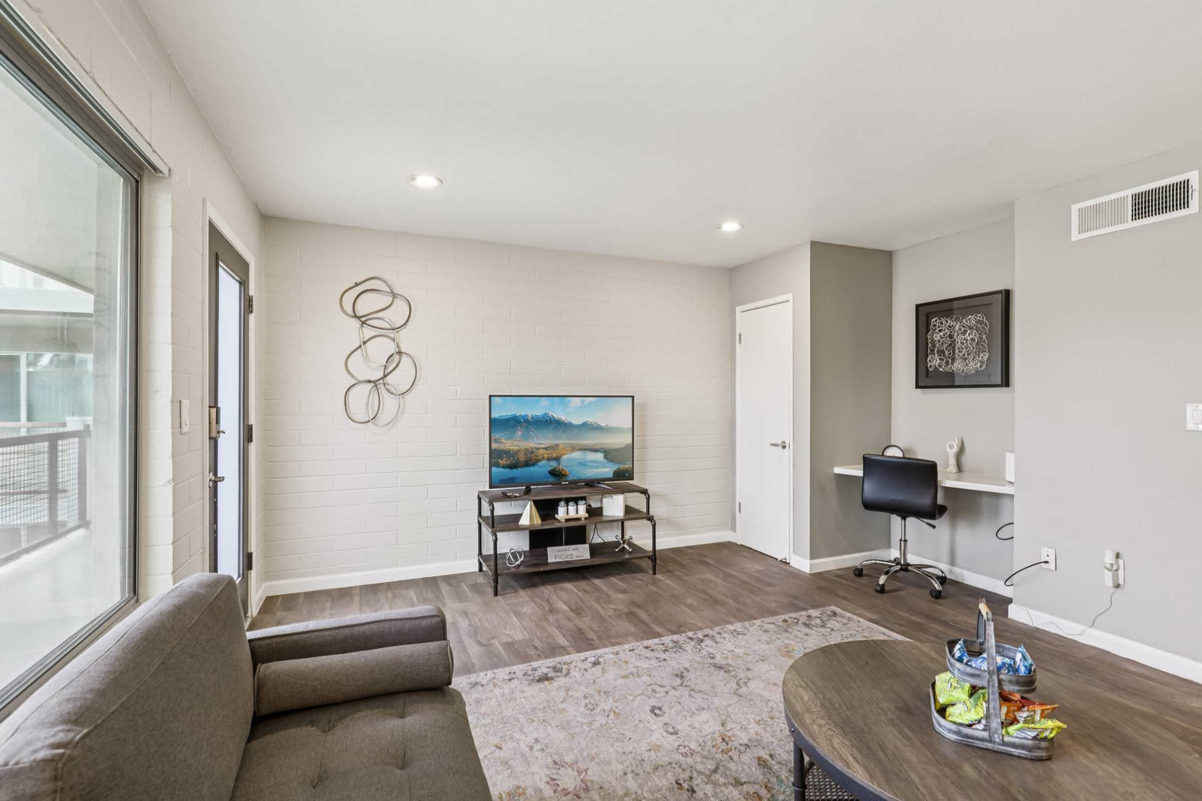 A modern living room featuring a grey sofa, wall-mounted TV displaying a scenic landscape, a round coffee table with snacks and drinks, and a desk with a chair. The walls are light-colored, and there's a door leading outside. Natural light enters through large windows, creating a bright and inviting atmosphere.