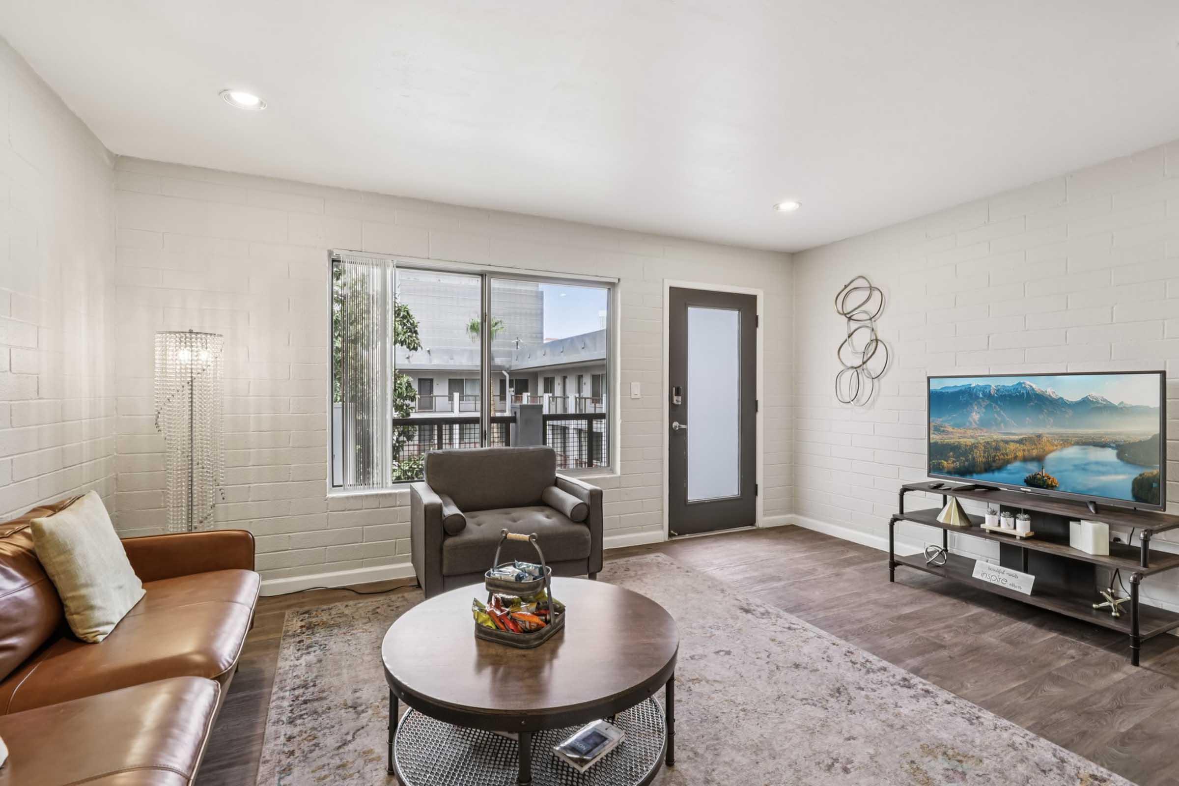 Cozy living room featuring a brown leather sofa, a round coffee table with decorative items, and a modern TV stand with a television displaying a scenic mountain view. A large window provides natural light, and a door leads outside. Wall art and ambient lighting enhance the inviting atmosphere.