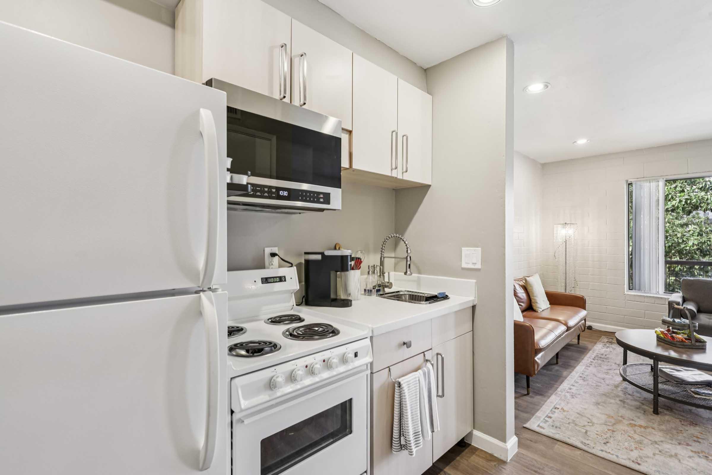 Modern kitchen featuring white appliances, including a refrigerator, stove, and microwave. The countertops are light-colored, and there is a coffee maker on the counter. In the background, a cozy living area is visible with a brown sofa and a round coffee table, creating a warm and inviting atmosphere.