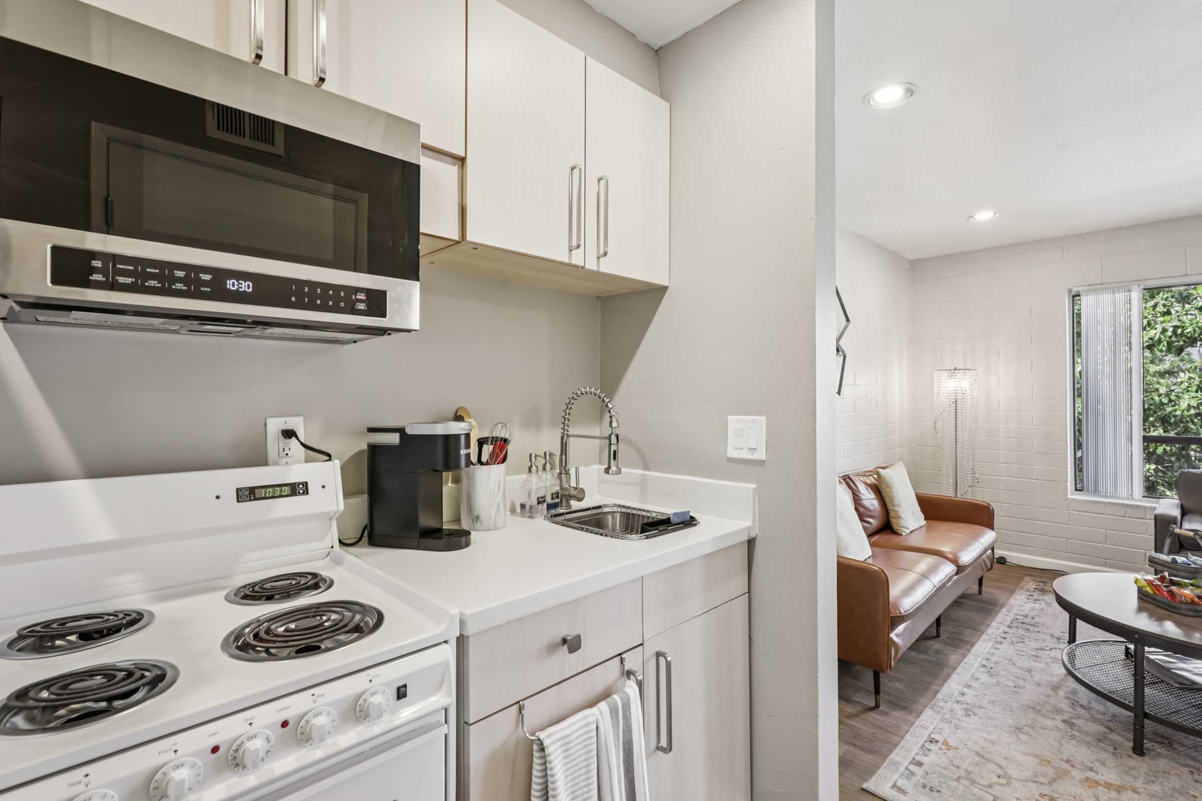 A modern kitchen featuring a stovetop, microwave, sink, and coffee maker. The kitchen has light-colored cabinets and is connected to a cozy living area with a sofa, a rug, and a window allowing natural light. The decor is contemporary and inviting.