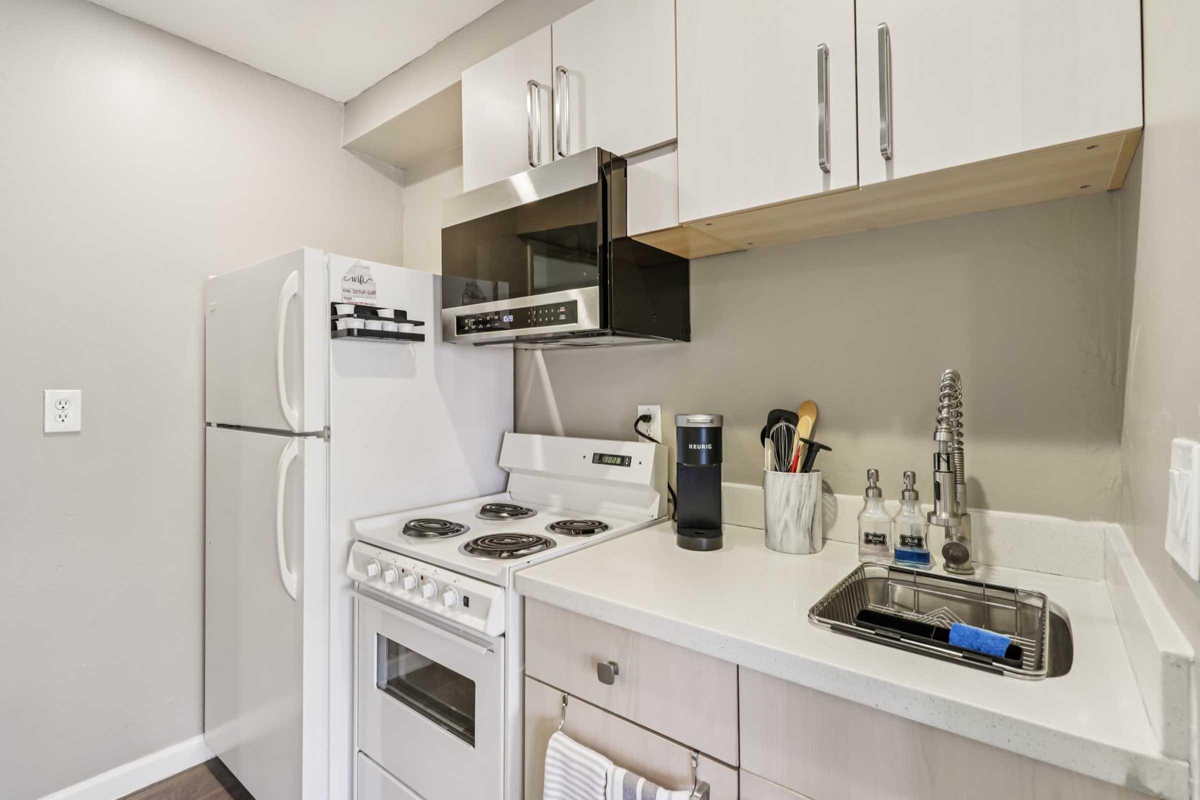 A modern kitchen featuring a white refrigerator, microwave, and stove. The countertops are light-colored with a sink that has a faucet. Kitchen tools and condiments are arranged neatly on the counter, along with a coffee maker. The cabinetry above is minimalistic and stylish.