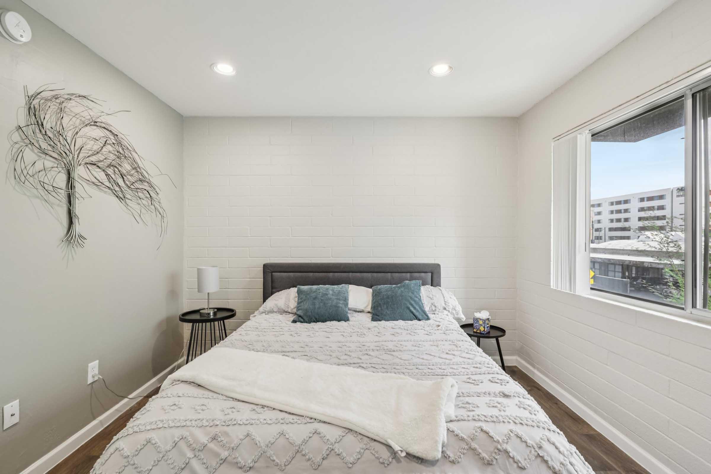 Modern bedroom featuring a gray accent wall with a decorative wall art made of thin branches. A neatly made bed with a light comforter and blue cushions is centered in the room. There are two side tables, one with a lamp and the other with a small decorative item, and a large window providing natural light.