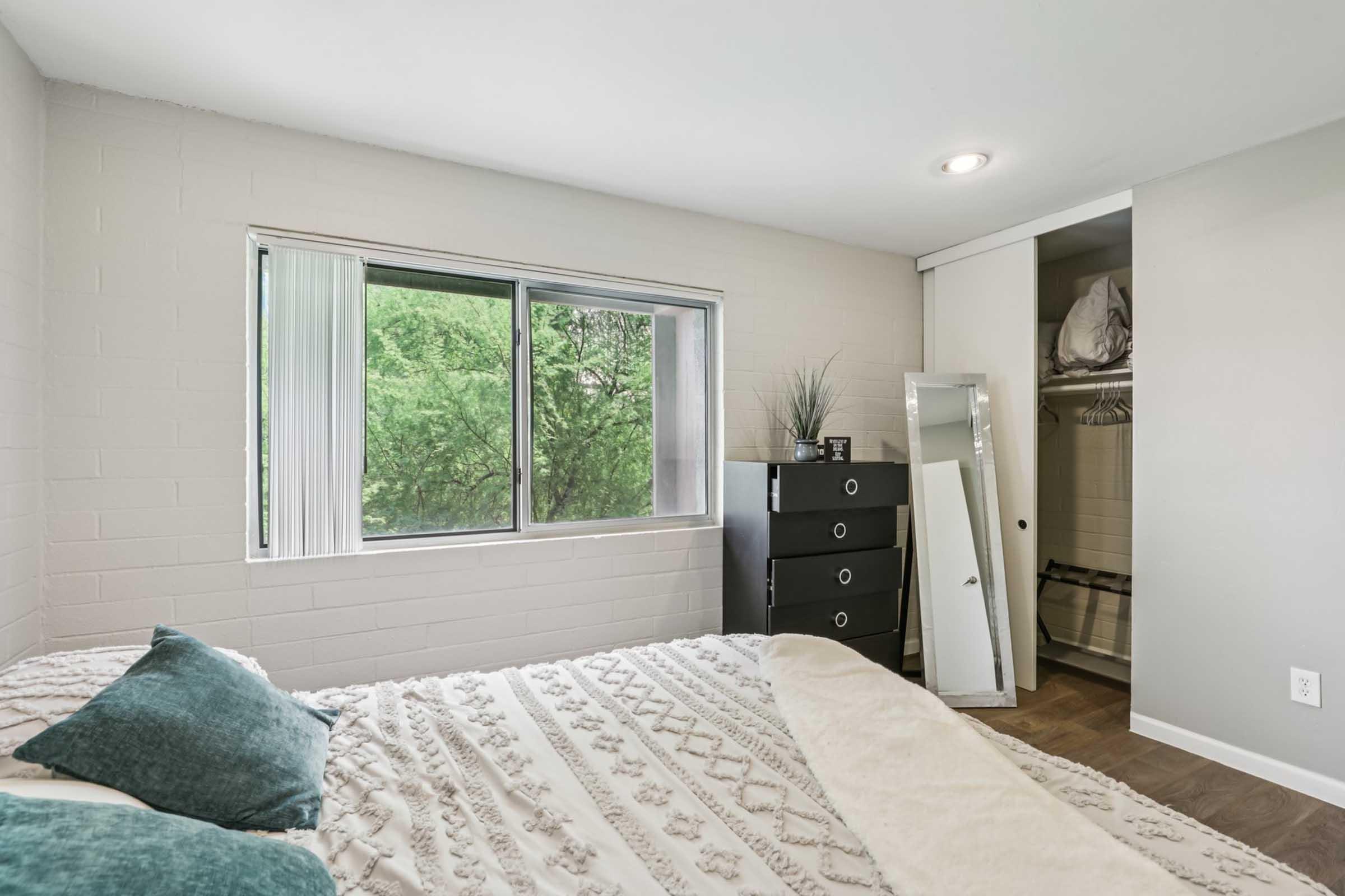 A cozy bedroom featuring a bed with decorative pillows, a large window showcasing greenery outside, a black dresser, a full-length mirror, and a closet with sliding doors. The walls are painted in light colors, creating a bright and inviting atmosphere.