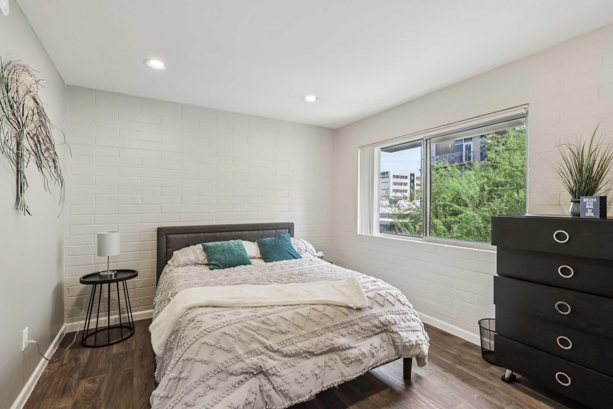 A modern bedroom featuring a neatly made bed with a gray headboard and white bedding. There is a small round side table with a lamp next to the bed. A large window allows natural light to enter, showcasing greenery outside. A black dresser with drawers is positioned against the wall, adding to the minimalist decor.
