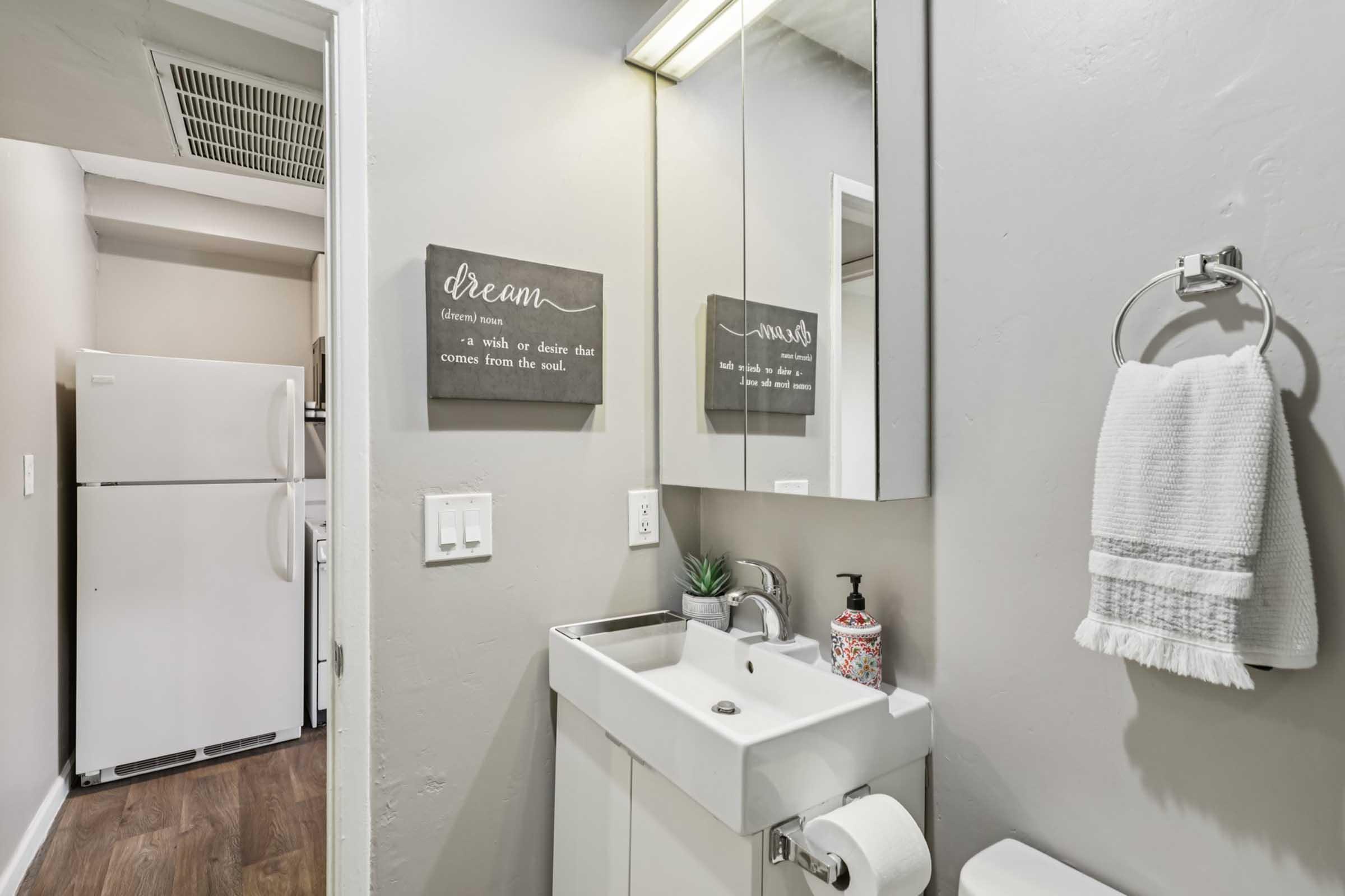 A small, modern bathroom featuring a white sink and a mirror. The walls are painted light gray, and there are inspirational quotes framed on the wall. A towel hangs nearby on a ring, and a small plant adds a decorative touch. In the background, a white refrigerator is visible in a separate area.