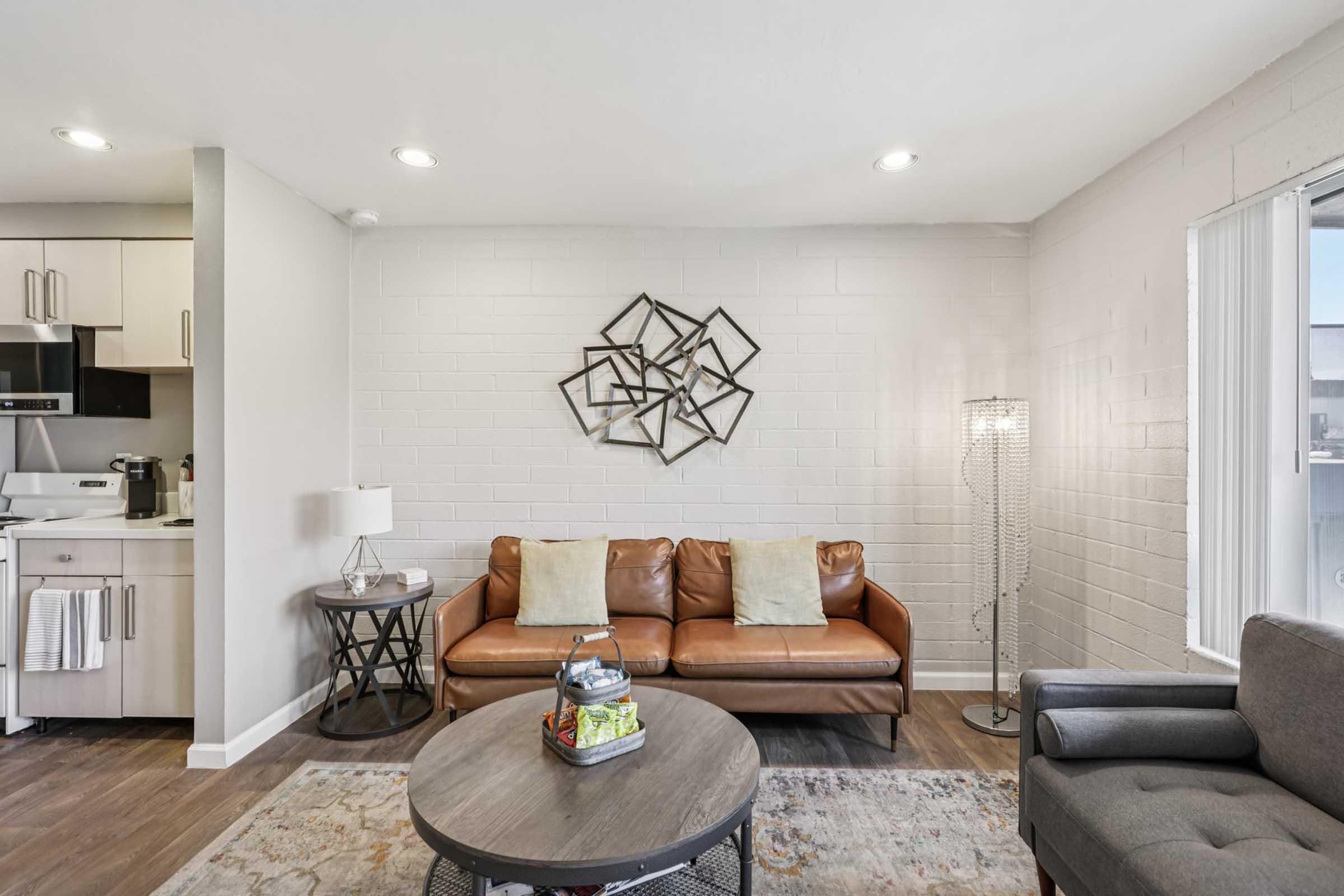 A modern living room featuring a brown leather couch with two pillows, a gray armchair, a round coffee table with a decorative item, and a geometric wall art piece. The space is well-lit with natural light coming from a window, and a floor lamp stands next to the couch. A kitchen area is visible in the background.