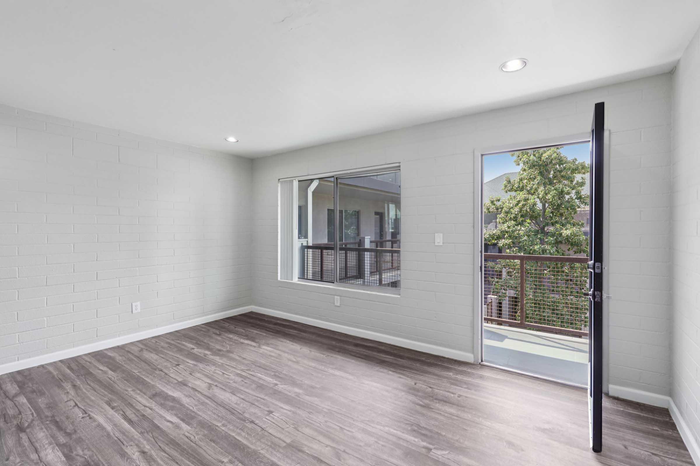 Empty room with light gray walls and wood-like flooring. There is a large window allowing natural light and a door leading to a balcony or outdoor area. The space is open and minimalist, suitable for personal decoration.