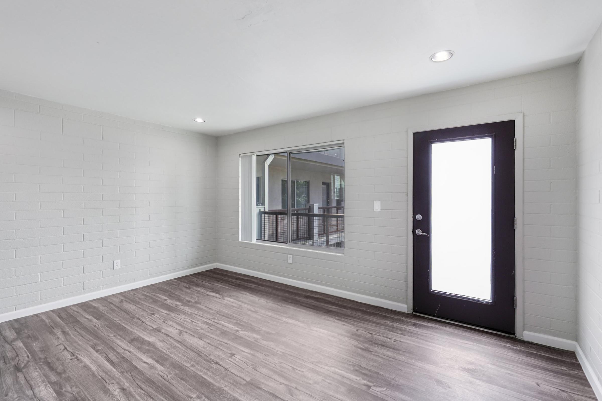 A spacious, empty room featuring light gray walls and a large window. The floor is made of dark laminate. A modern black front door is visible, leading outside. The room is well-lit with natural light coming through the window.