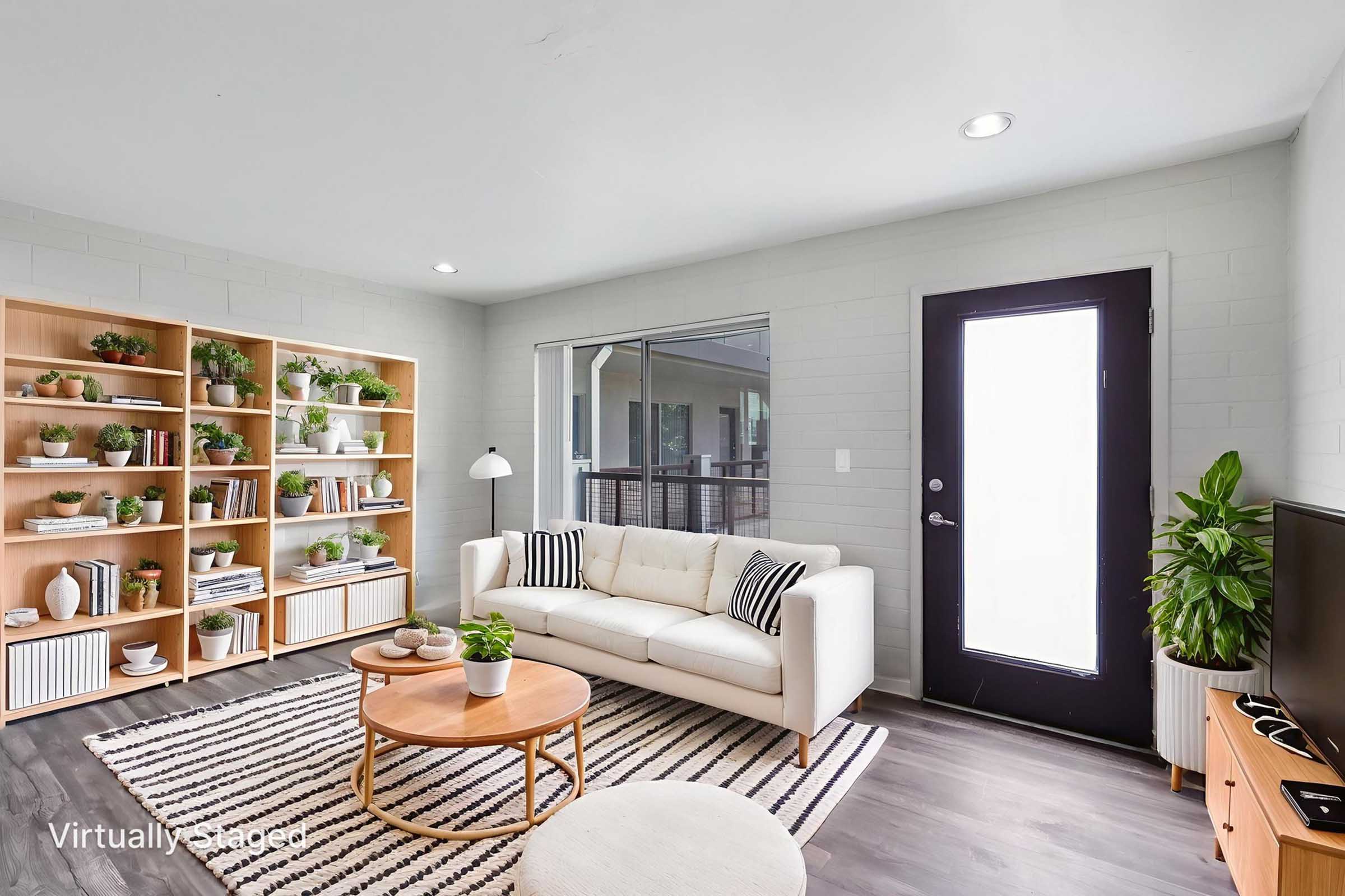 A modern living room featuring a light-colored sofa, a round wooden coffee table, and a large bookshelf filled with plants and decorative items. Natural light enters through a glass door leading to a small balcony. The room has a neutral color palette with striped rug flooring.