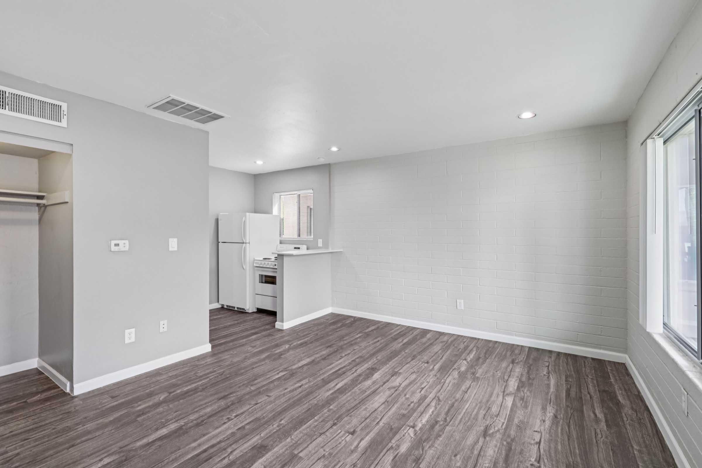 A modern apartment interior featuring an open layout with light gray walls and hardwood-style flooring. The space includes a white refrigerator and kitchen area with a bar. Natural light enters through a large window, and there's a closet nook on one side of the room.