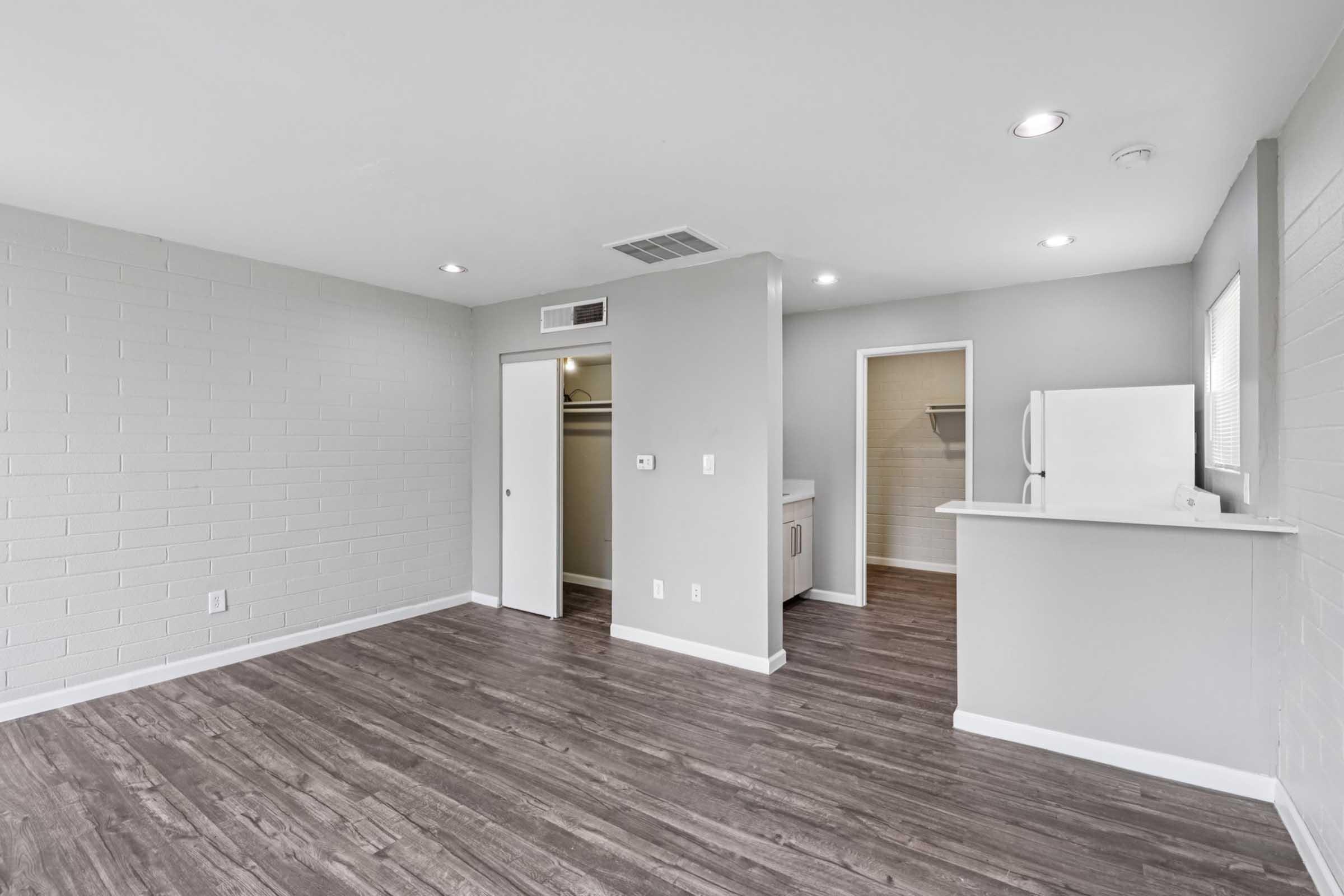 A spacious, modern living area with light gray walls and dark wood-like flooring. The room features recessed lighting, a doorway leading to a closet, and an open area with a kitchenette visible in the background, including a white refrigerator and cabinets. Natural light enters through a nearby window.