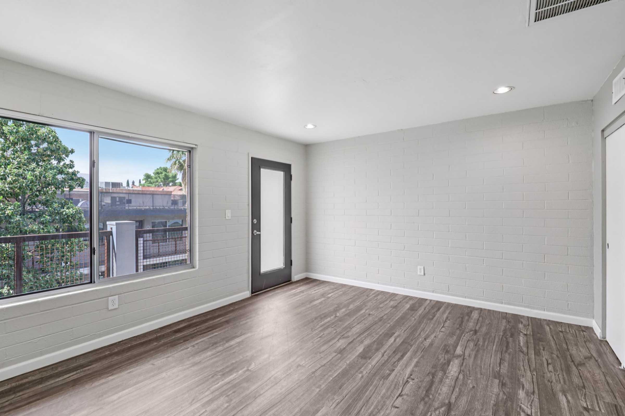 A bright, empty room with light gray walls and modern wood-style flooring. A large window allows natural light to flood the space, offering a view of greenery outside. A door leads to a balcony, and there are recessed lights in the ceiling, creating a clean and open atmosphere.