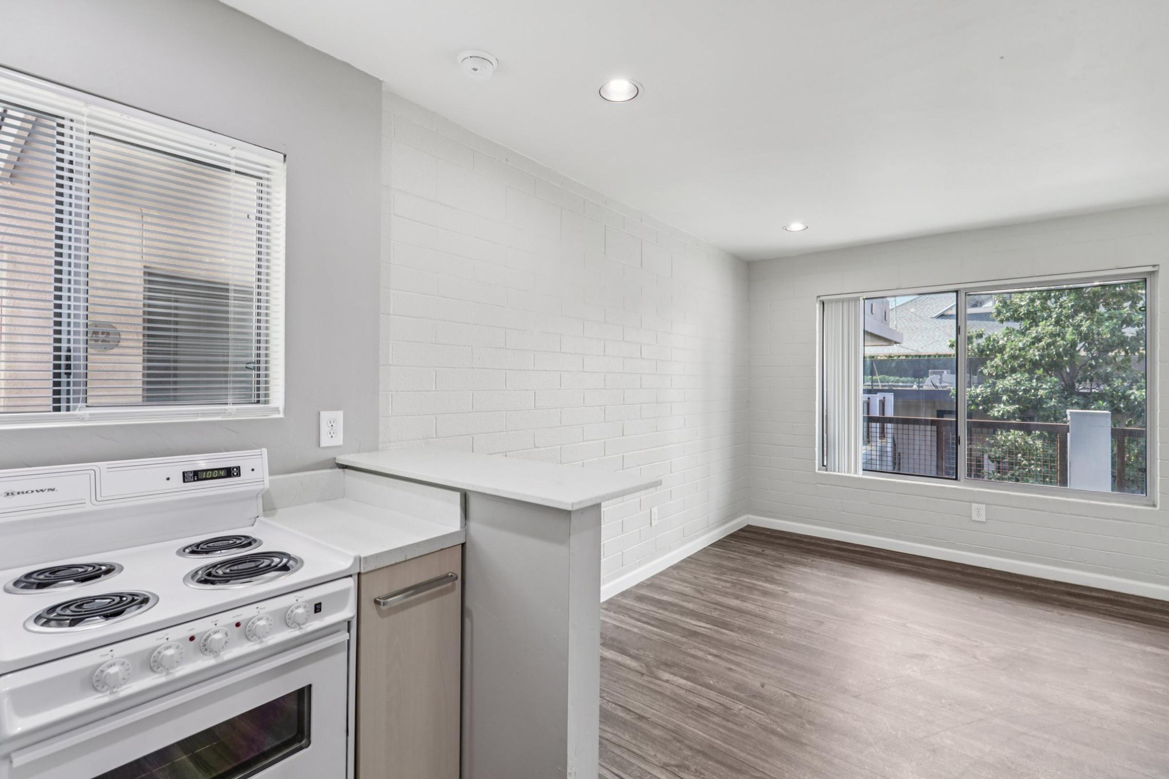 A modern kitchen space featuring a white stove, a sleek countertop, and a window with blinds. The adjacent living area has clean, light-colored walls and a large window overlooking greenery outside. The flooring is a warm wood laminate, creating a bright and inviting atmosphere.