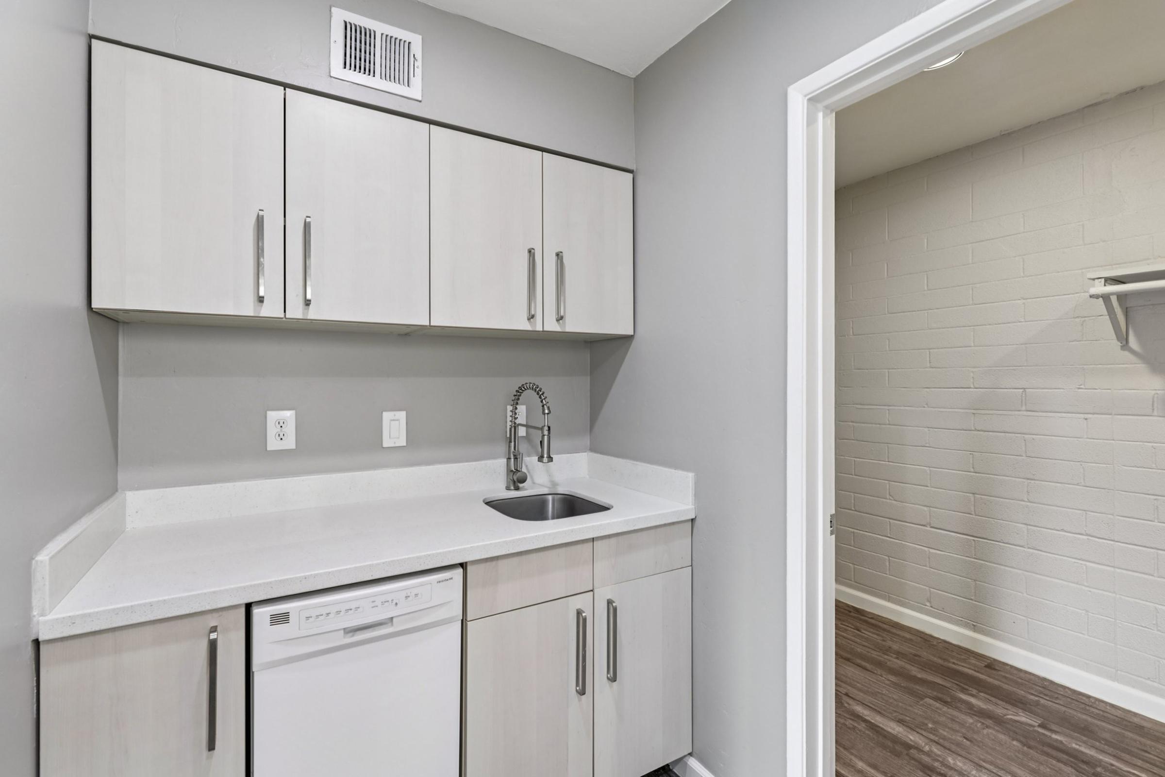 Modern kitchen space featuring light gray cabinets, a countertop with a stainless steel sink and faucet, and a built-in dishwasher. The design includes a small area adjacent to a closet or storage space, with light-colored walls and a wood-like floor.